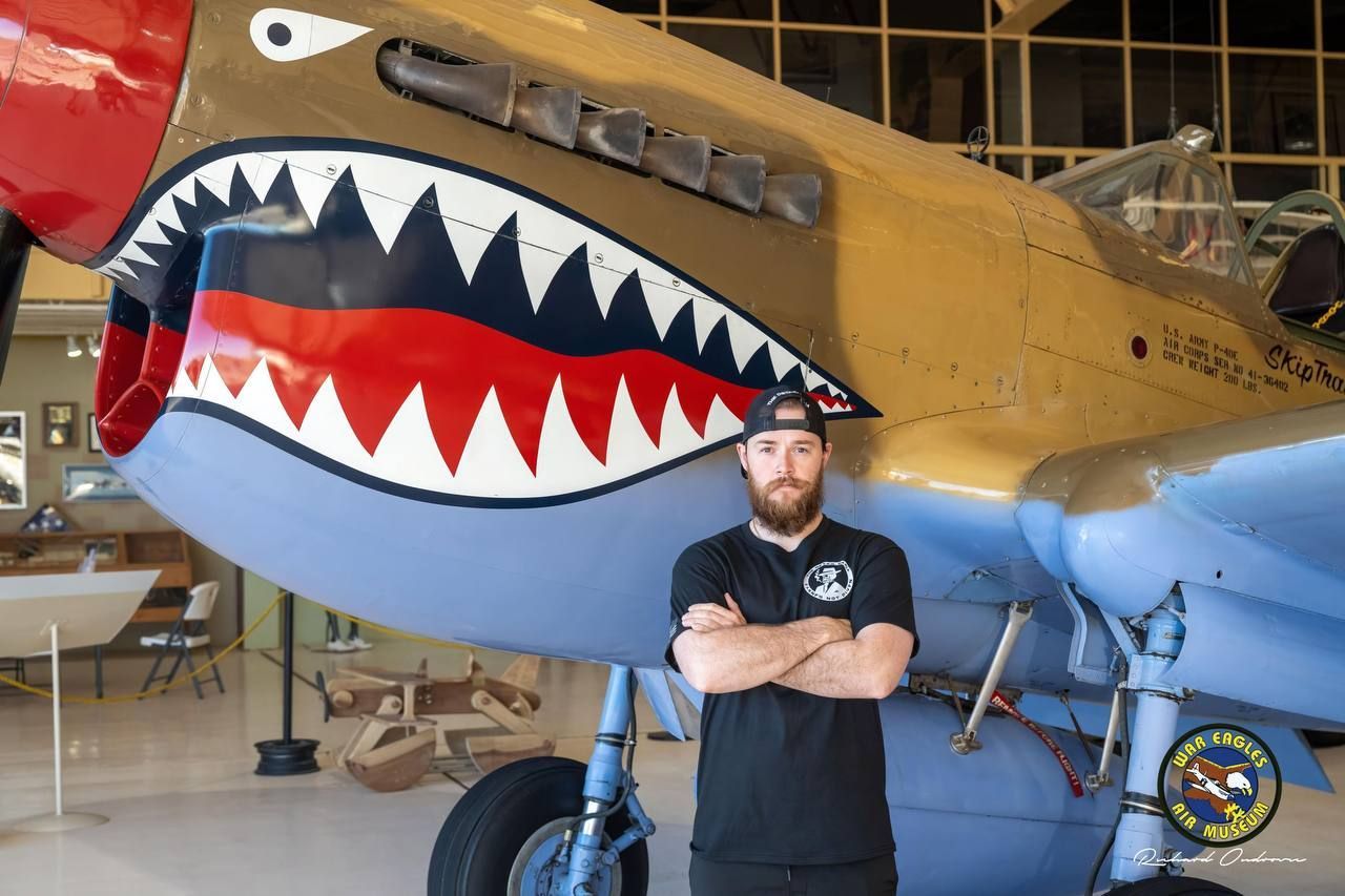 Man with crossed arms stands in front of a P-40 Warhawk fighter plane with shark mouth, inside a hangar.