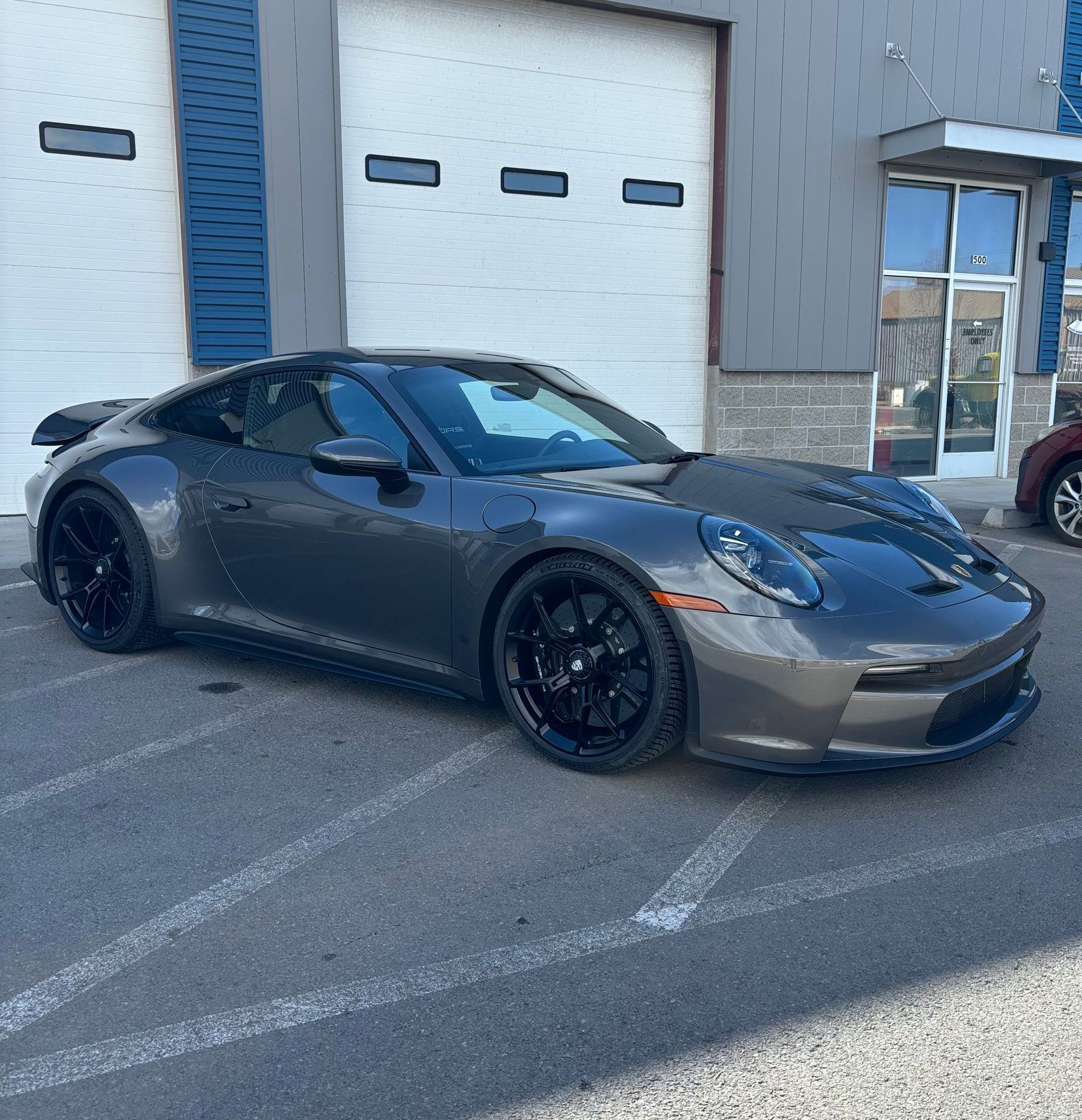 Dark grey Porsche 911 parked in front of a building with white garage doors and a blue exterior wall. Black wheels.