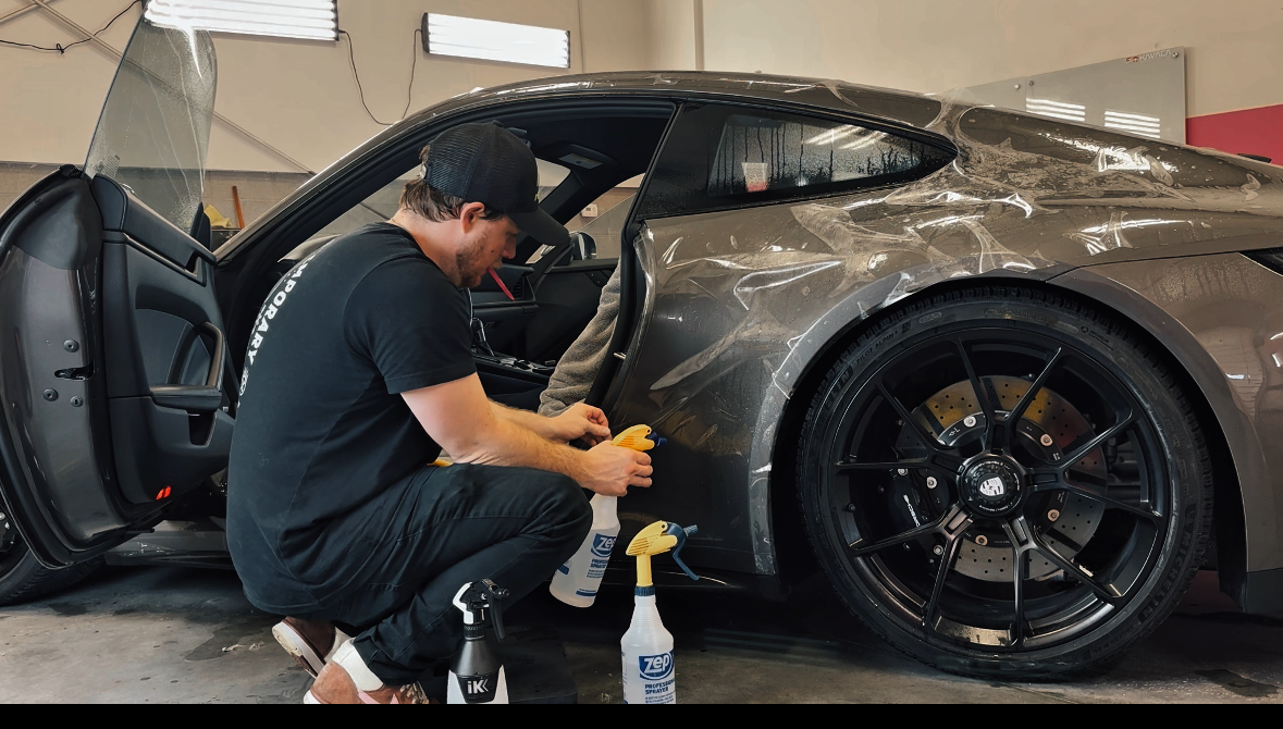 A man applies protective film to a gray sports car in a garage.