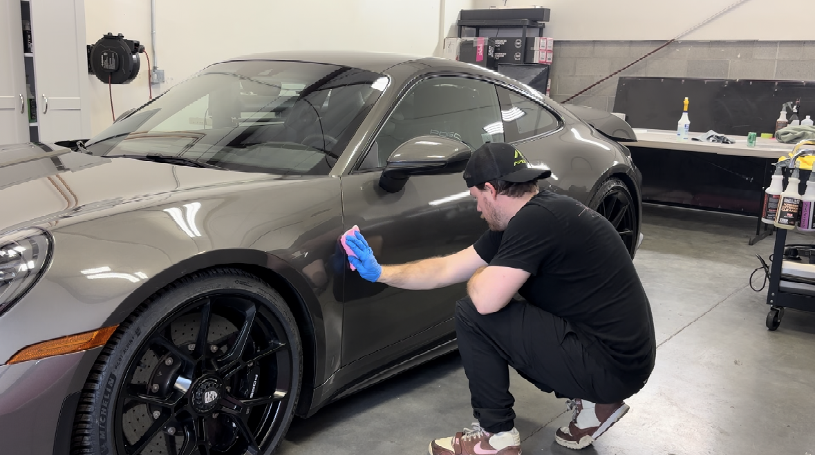 Man wearing a cap and gloves, cleaning a gray sports car in a garage. He is squatting and wiping the car's side.