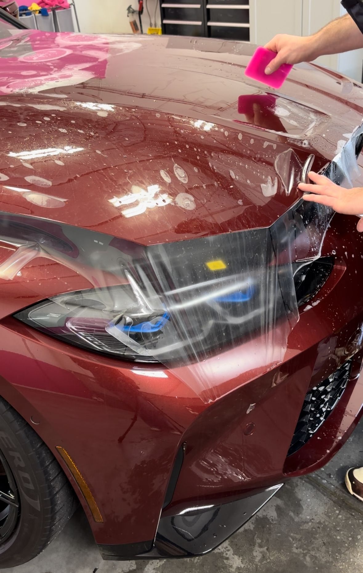 Applying clear protective film to a maroon car's hood, using a squeegee to smooth out air bubbles.