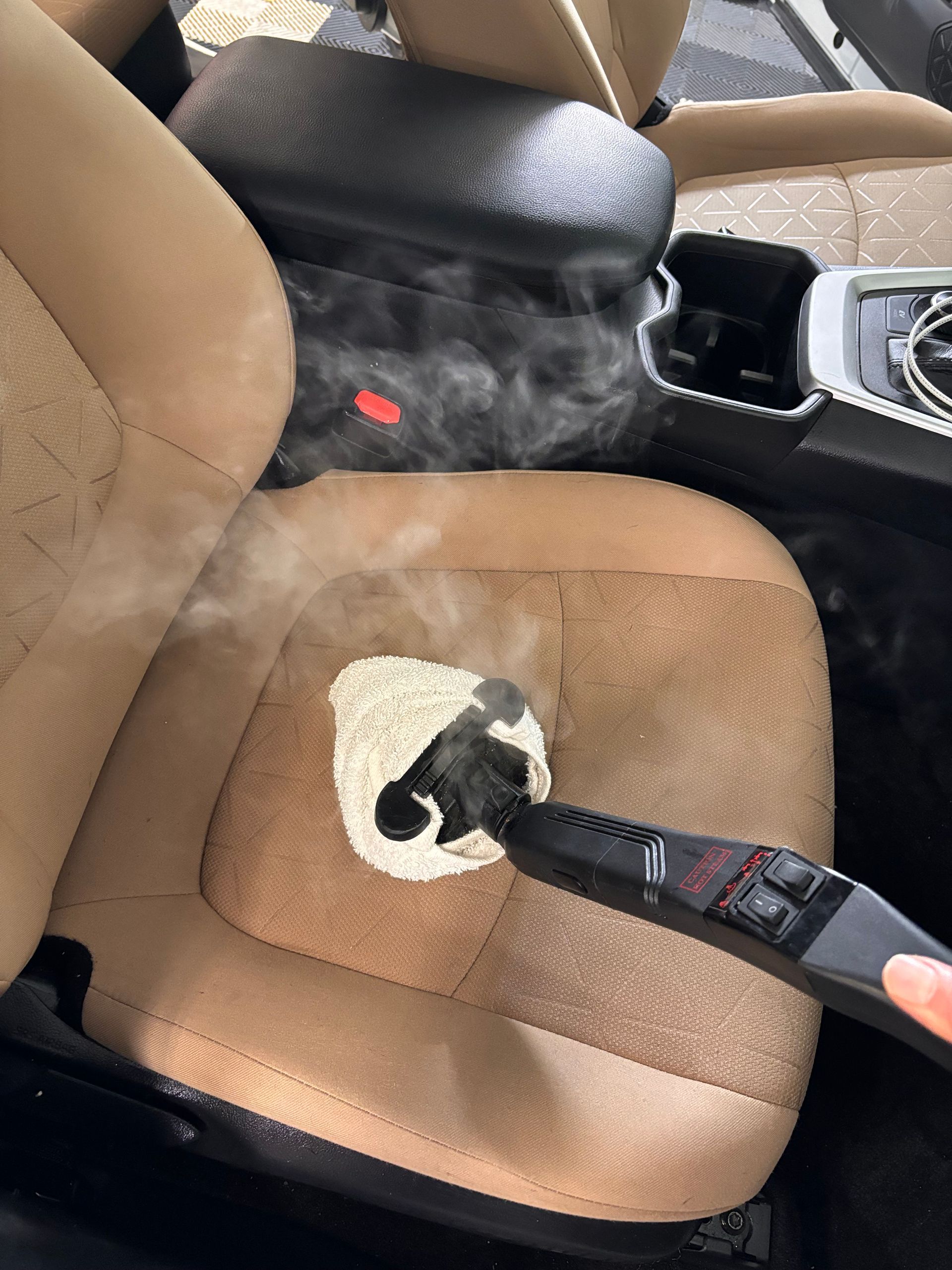 Steaming car seat; steam cleaner removes stain from tan upholstery inside a vehicle.