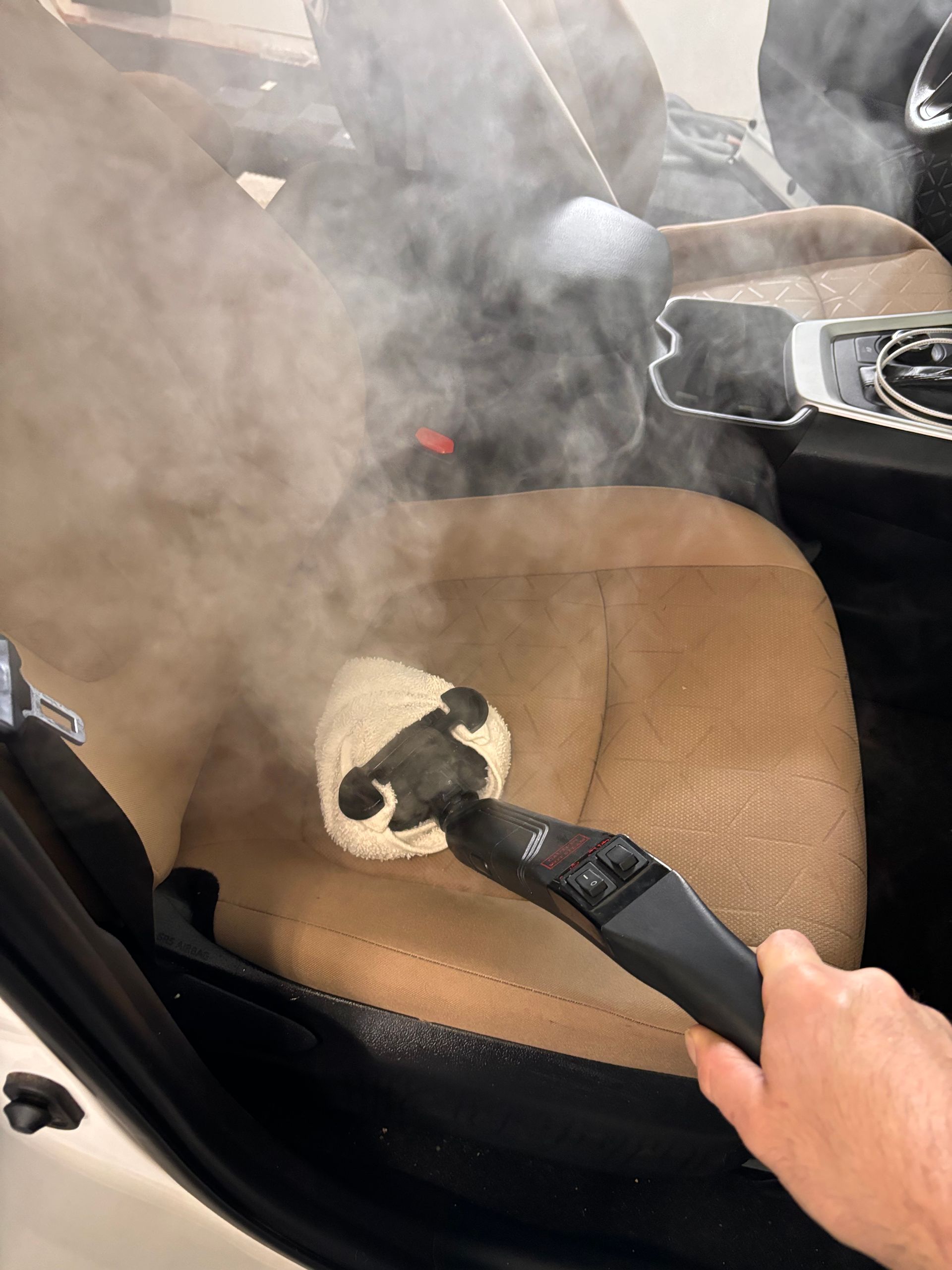 Steaming a tan car seat with a handheld steam cleaner, creating a cloud of vapor.