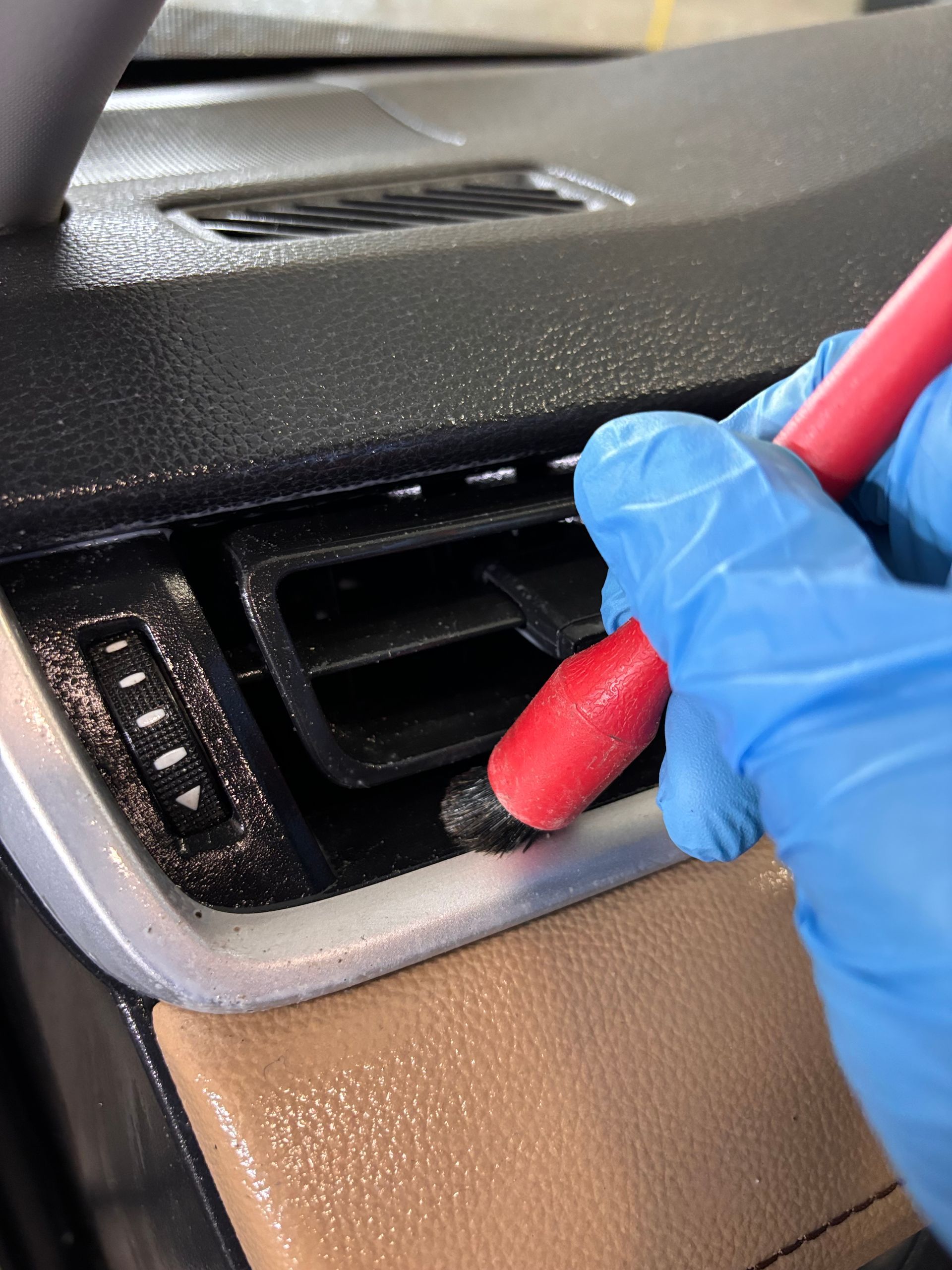 A gloved hand cleans a car's air vent with a red brush; dust is visible on the dashboard and vents.