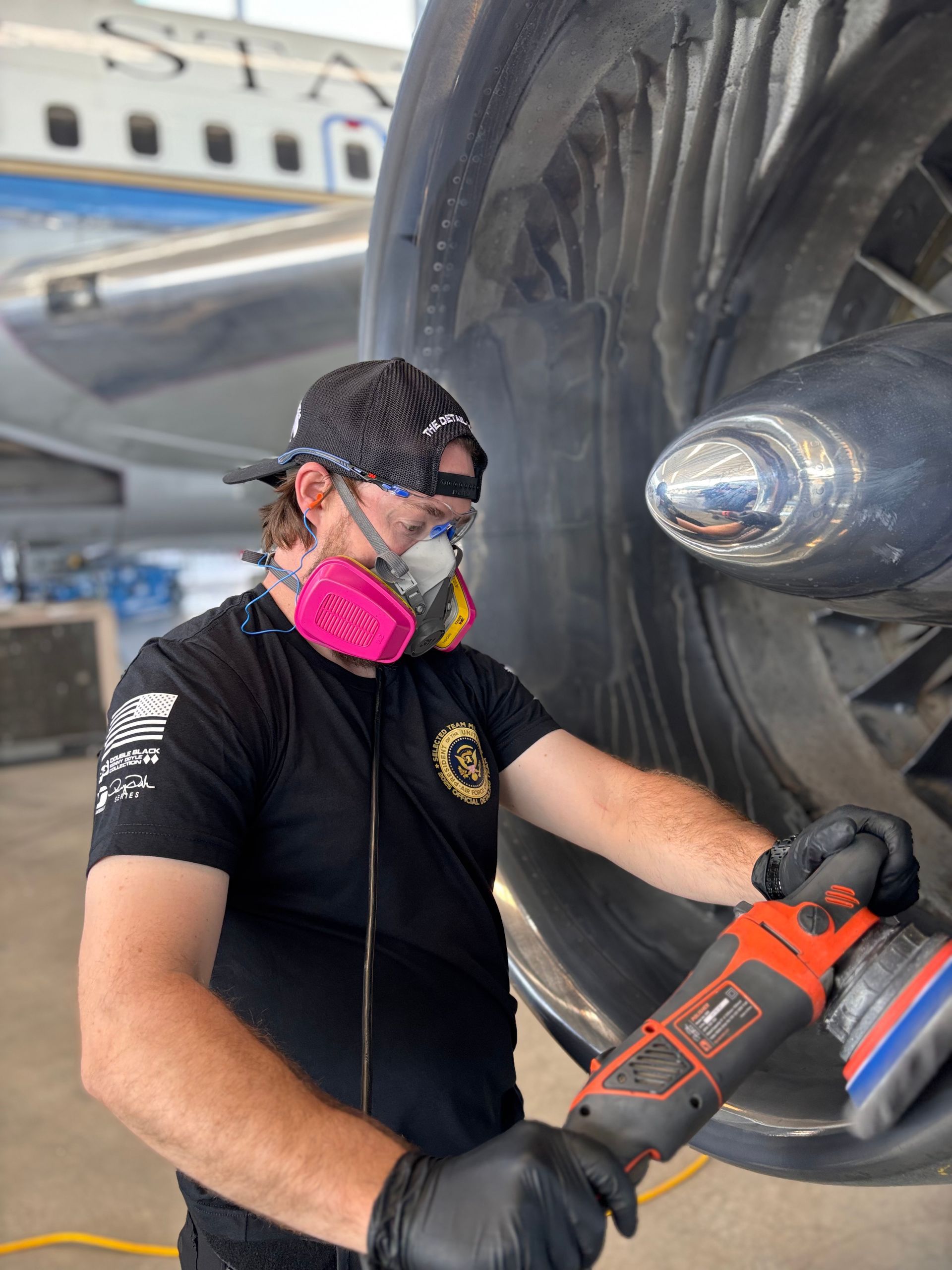 Man polishing an aircraft engine with an electric buffer, wearing a respirator, in a hangar.