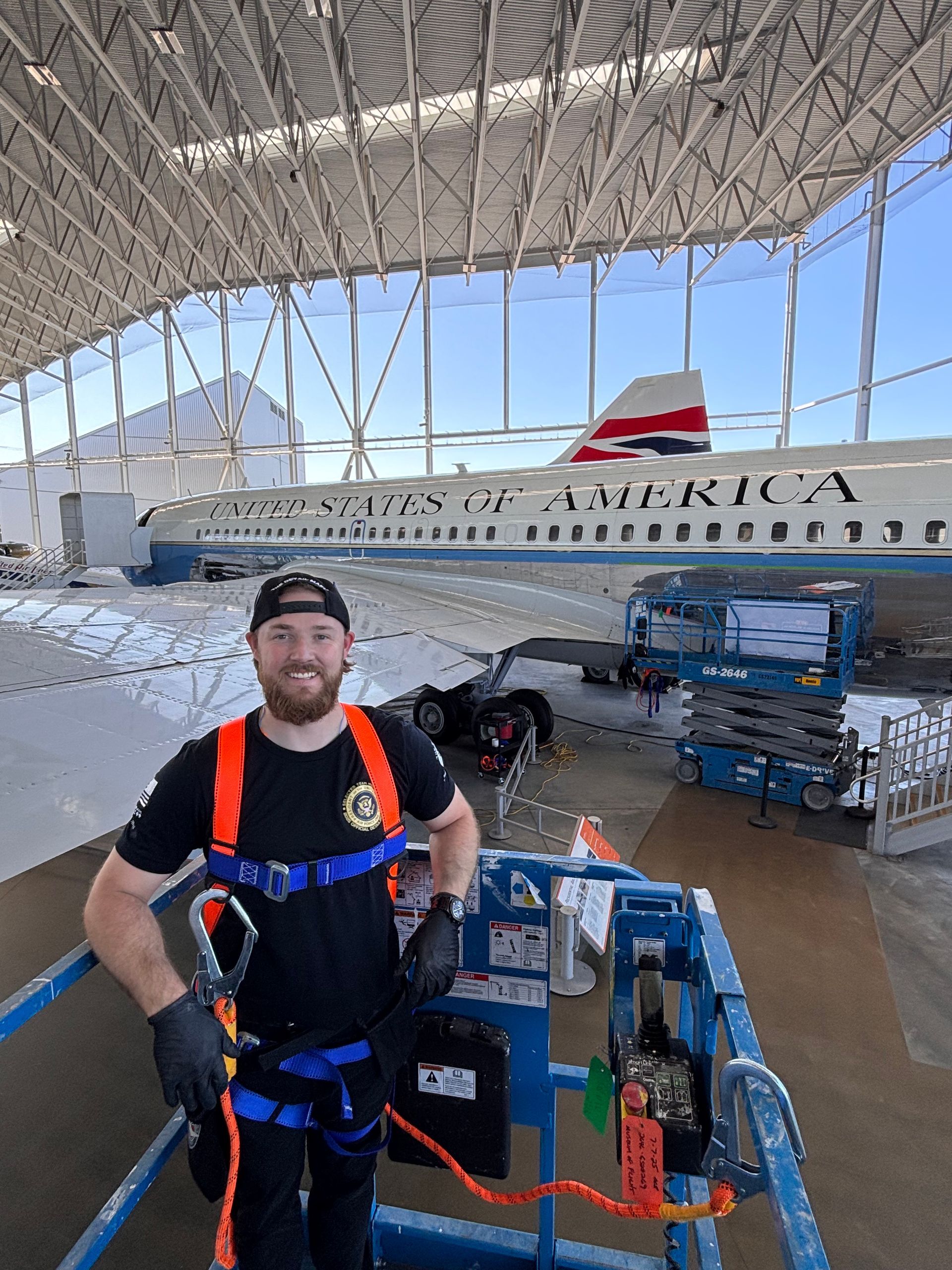Man in harness stands in front of a partially disassembled airplane in a hangar.