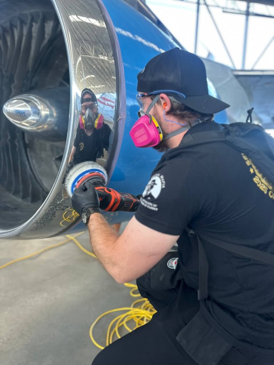 Person polishing a jet engine. They wear a mask, gloves, and a black shirt. The engine is blue and silver.