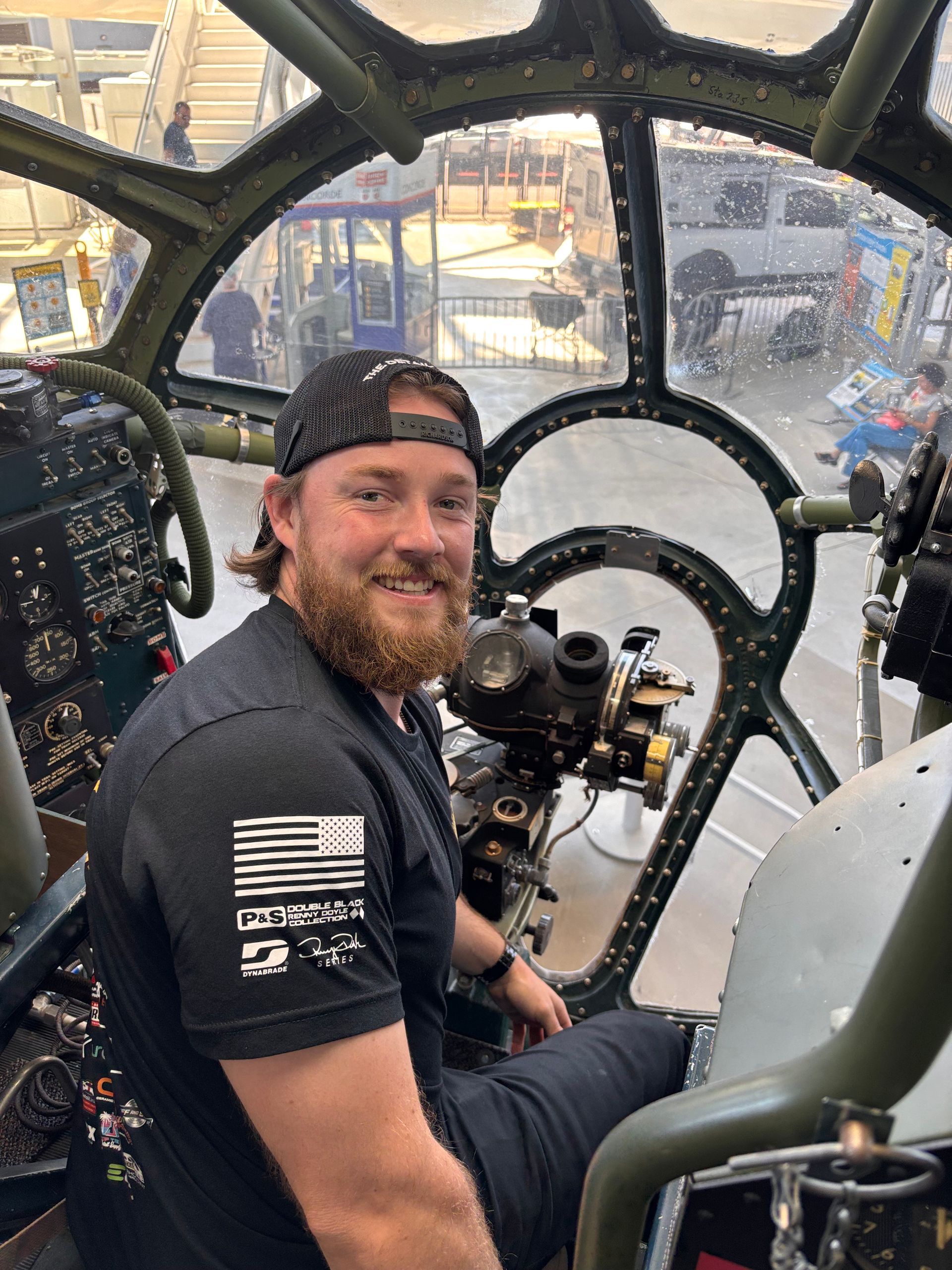 Man smiling in the cockpit of a vintage aircraft; black cap, short beard, dark shirt.