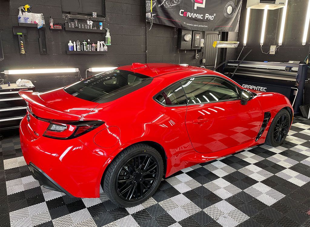 Red sports car in a garage with black rims, spoiler, and checkered floor.