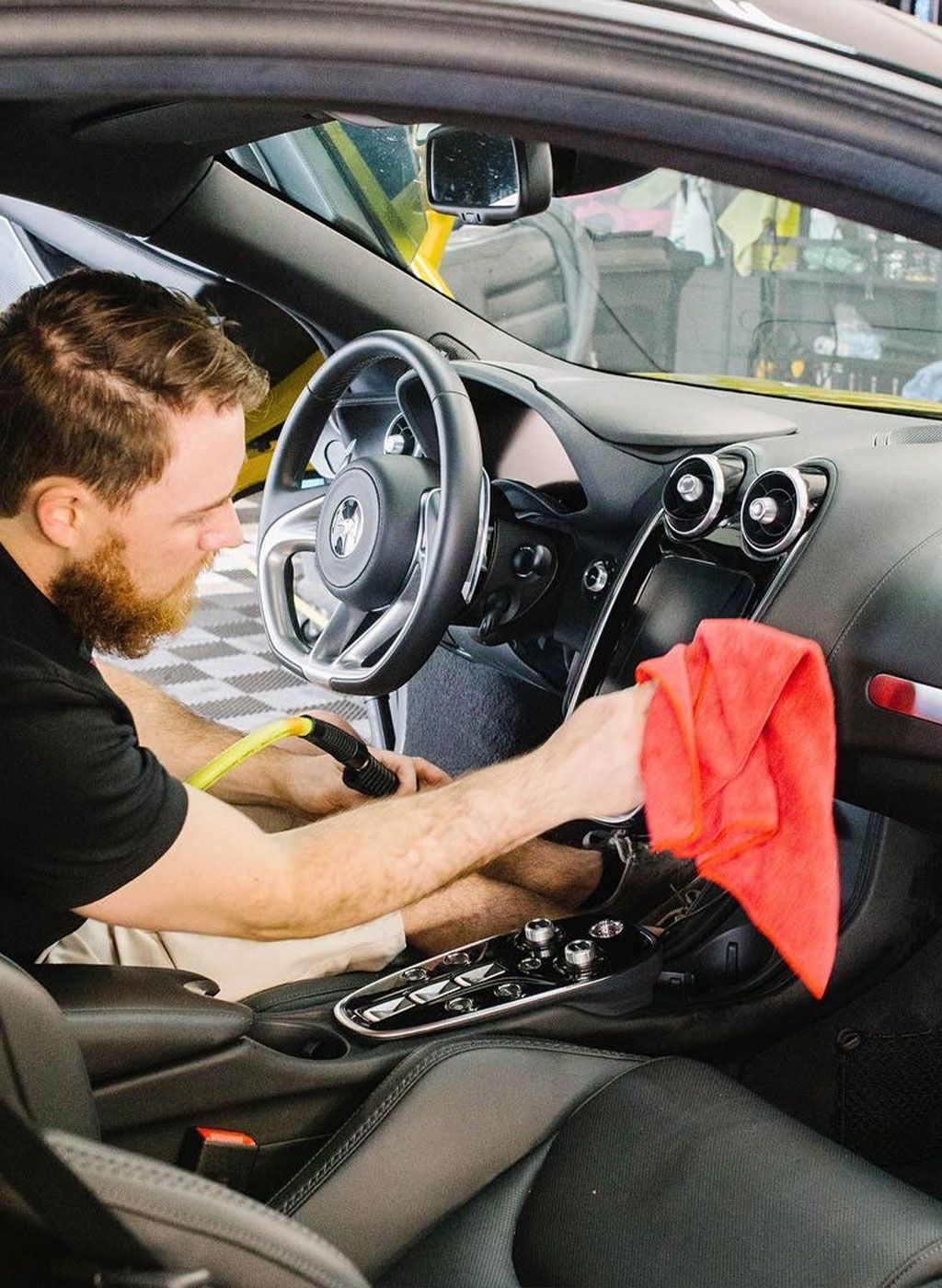 Man cleaning the dashboard of a yellow sports car with a red cloth and a detailing tool inside the vehicle.