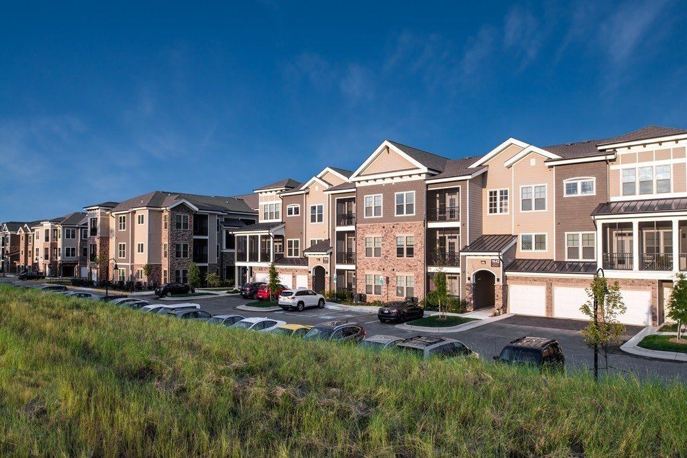 Apartment complex on a sunny day; multi-story buildings with tan, brown, and brick exteriors. Cars are parked in front, green grass in foreground.