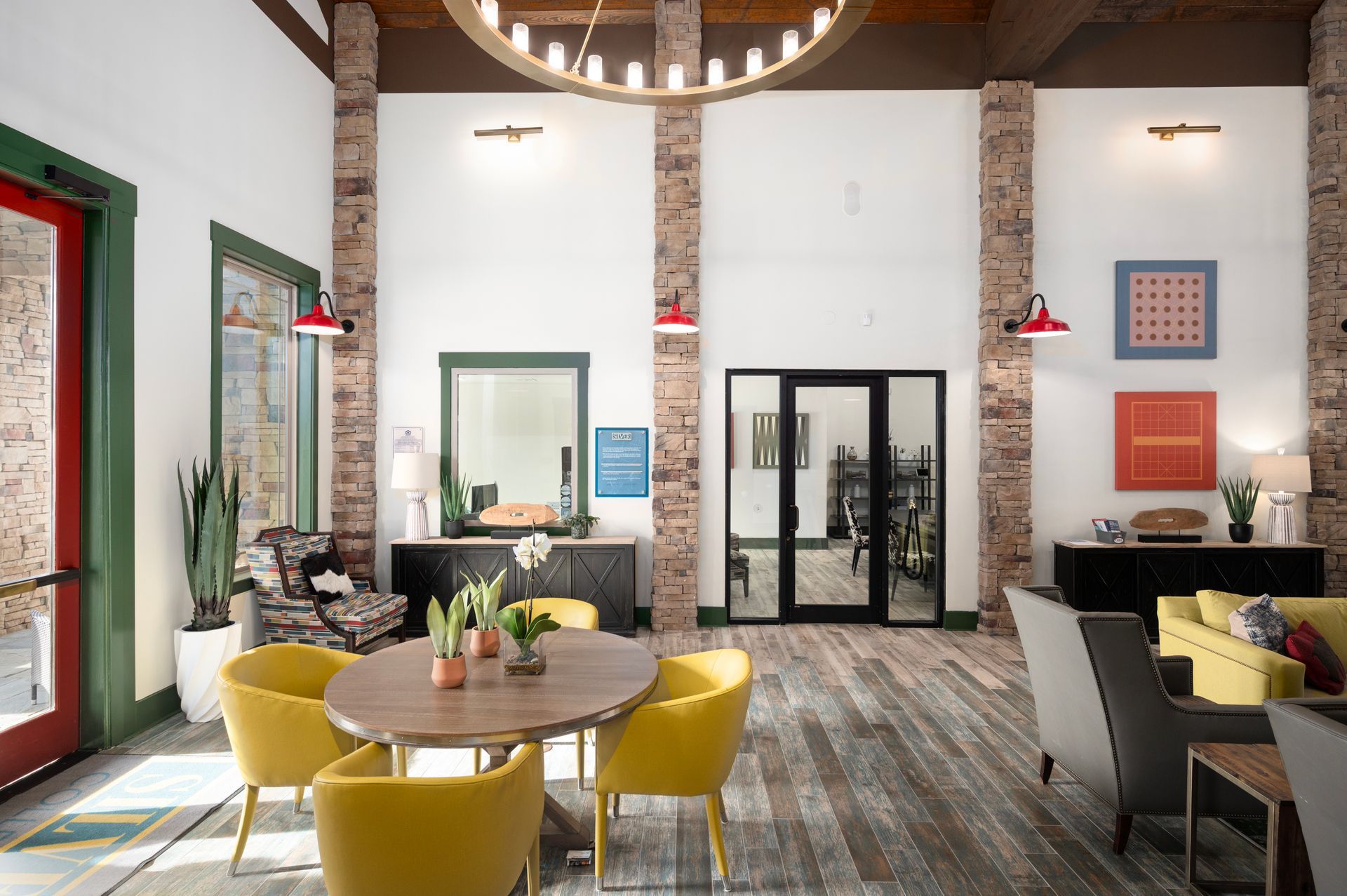 A hotel lobby with a round wooden table, yellow chairs, a lounge area, exposed brick columns, and white walls.
