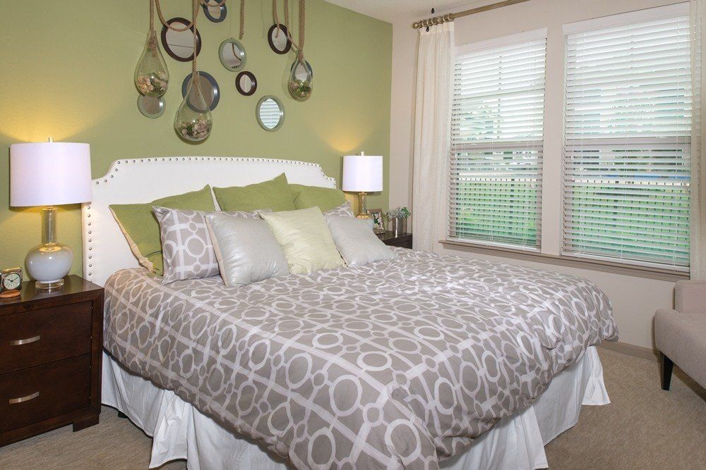 A bed with a white headboard, gray patterned bedding, and green accent pillows against a green wall with round mirrors at Silver Collection at Cosner's Corner in Fredericksburg, VA.