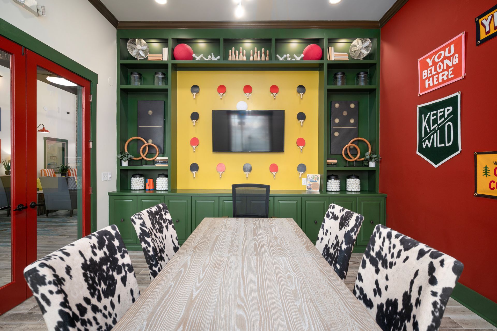 A conference room with a wooden table, cow-print chairs, a yellow wall with ping-pong paddles, and red accented walls at Silver Collection at Cosner's Corner in Fredericksburg, VA.