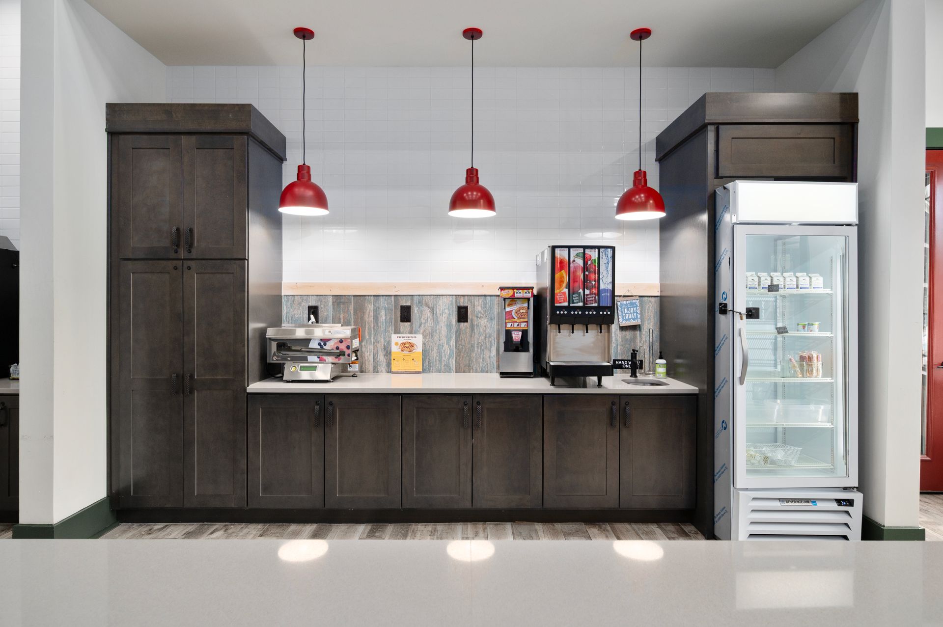A beverage and snack station with dark wooden cabinets, three red pendant lights, a soda fountain, and a glass fridge at Silver Collection at Cosner's Corner in Fredericksburg, VA.