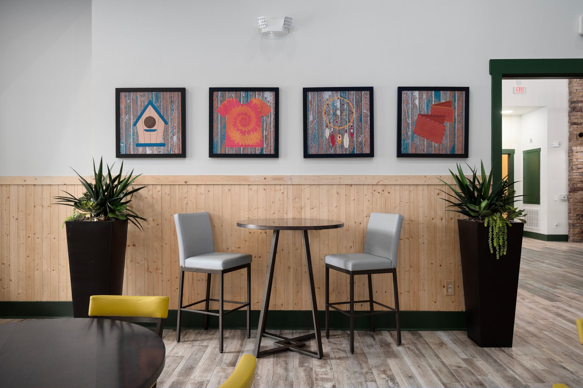 A high-top table with two grey chairs sits against a light wood-paneled wall decorated with four framed prints and plants at Silver Collection at Cosner's Corner in Fredericksburg, VA.