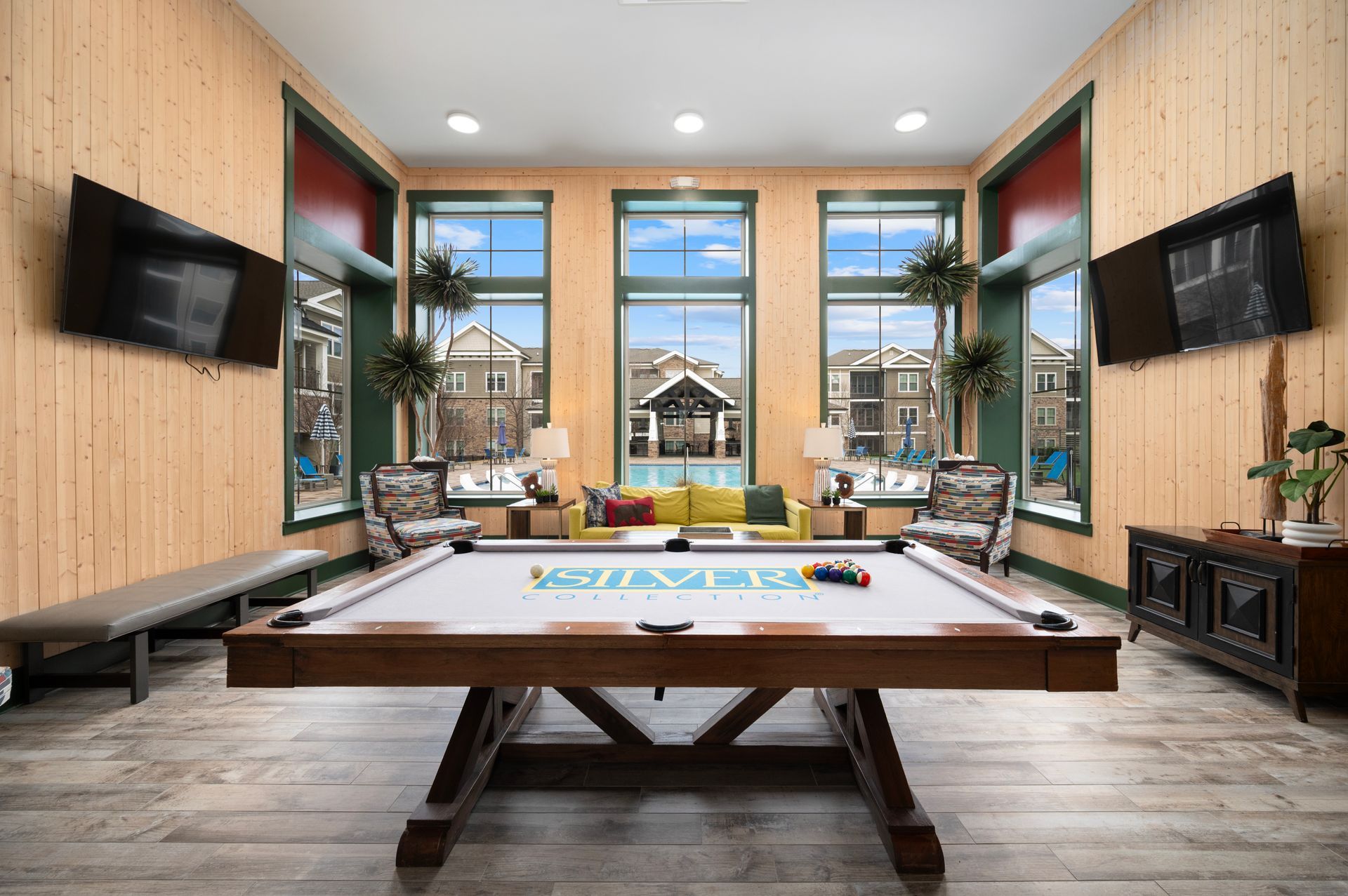 A pool table sits in the foreground of a bright community room with wood-paneled walls, seating areas, and large windows at Silver Collection at Cosner's Corner in Fredericksburg, VA.