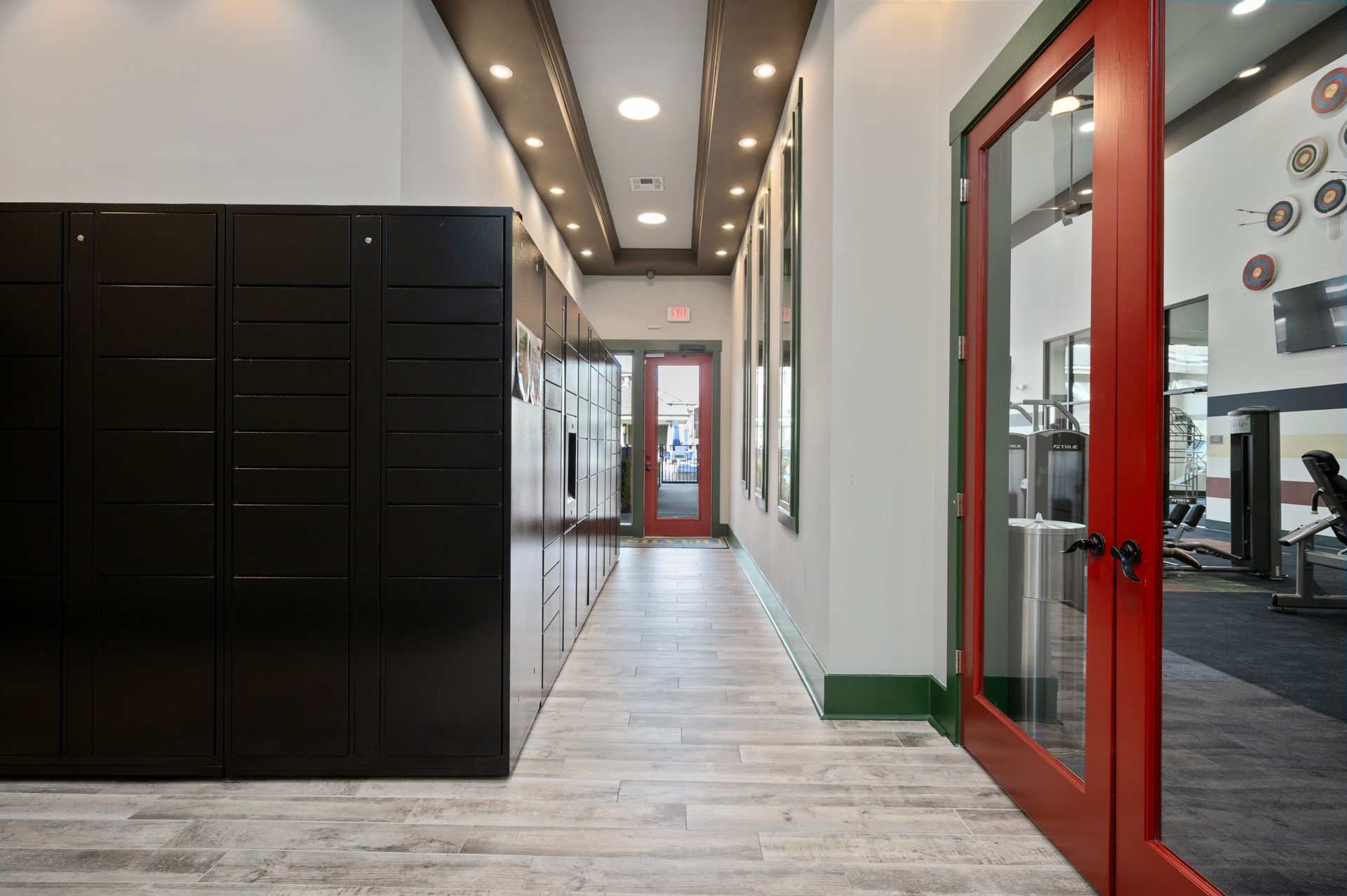A hallway with black storage lockers on the left, wood-look flooring, and a red-framed glass door leading to a gym at Silver Collection at Cosner's Corner in Fredericksburg, VA.
