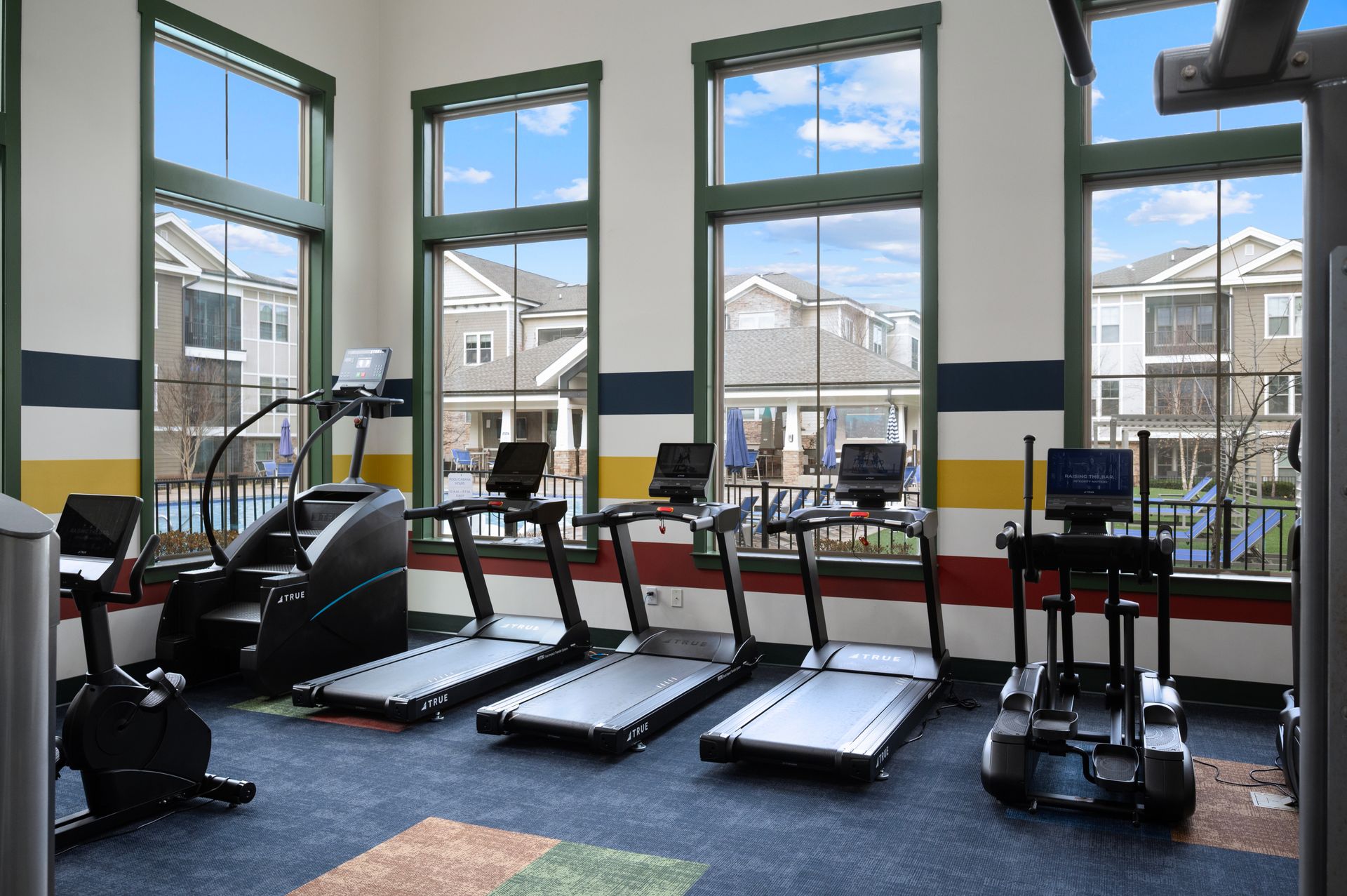 A fitness room with treadmills and cardio equipment lined up in front of large windows overlooking apartment buildings.