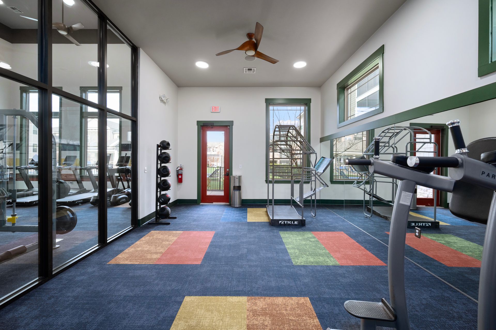 An indoor gym featuring a blue carpet with colored rectangular tiles, treadmills, exercise equipment, and a glass door at Silver Collection at Cosner's Corner in Fredericksburg, VA.