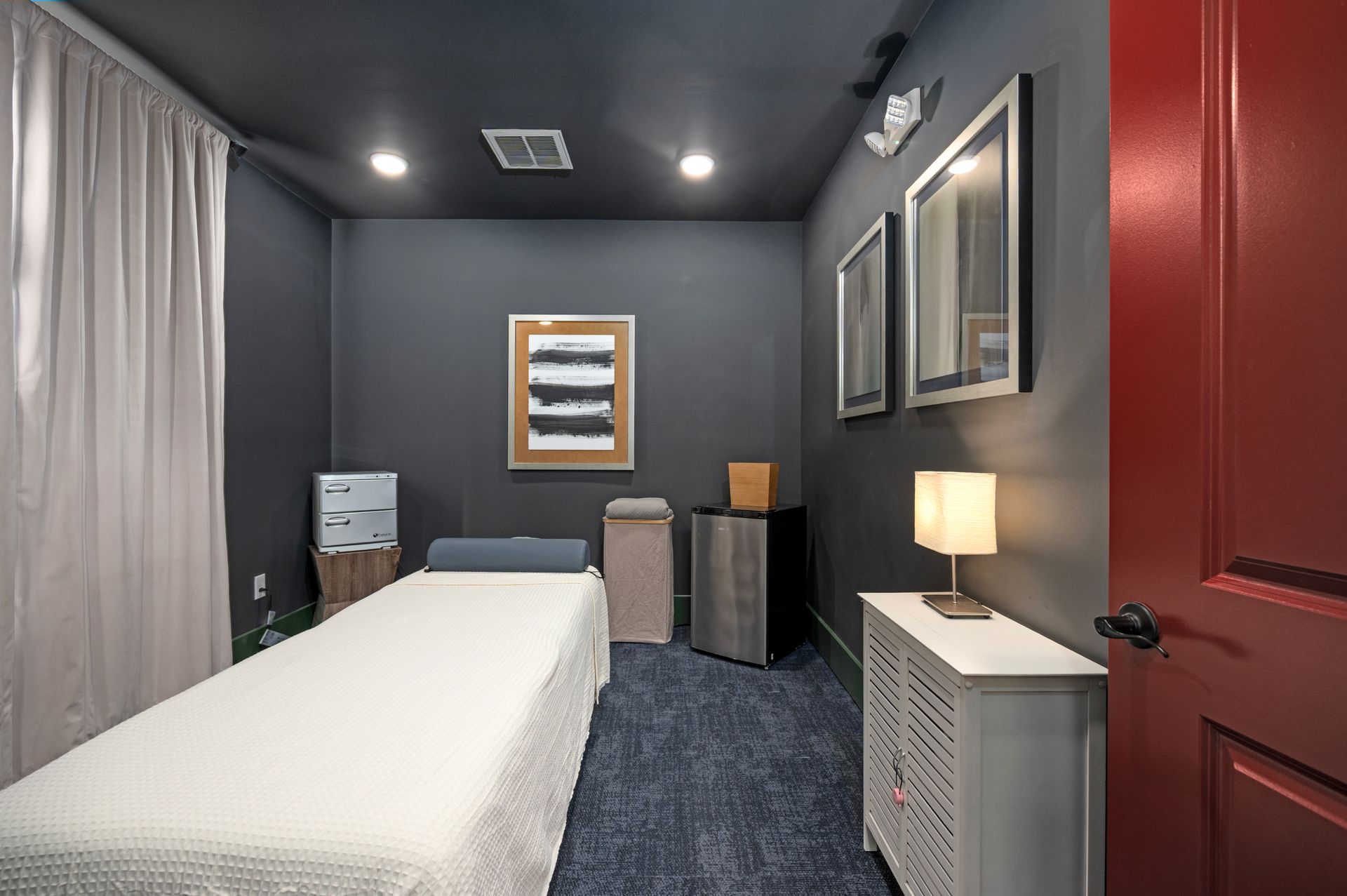 A treatment room with dark walls, a massage bed with white linens, a bedside table, and a bright red door at Silver Collection at Cosner's Corner in Fredericksburg, VA.
