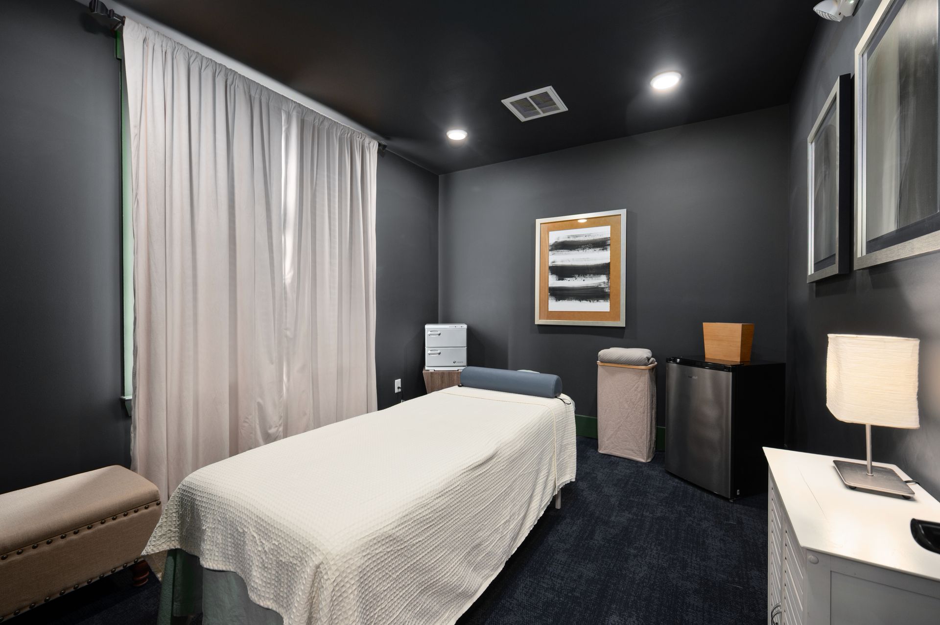 A massage room with dark gray walls, a treatment table, white curtains, and a small table with a lamp at Silver Collection at Cosner's Corner in Fredericksburg, VA.