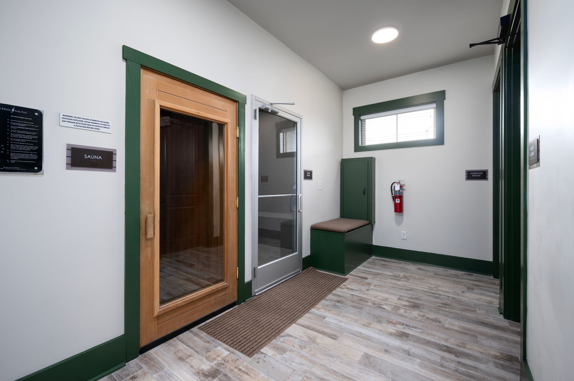 A room with a wooden sauna, a glass door, a green bench, a fire extinguisher, and light wood-grain flooring at Silver Collection at Cosner's Corner in Fredericksburg, VA.