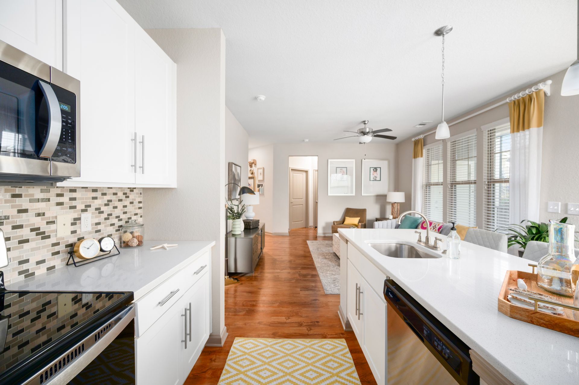 Modern white kitchen with stainless steel appliances, marble countertops, and a view into a living area with hardwood floors.