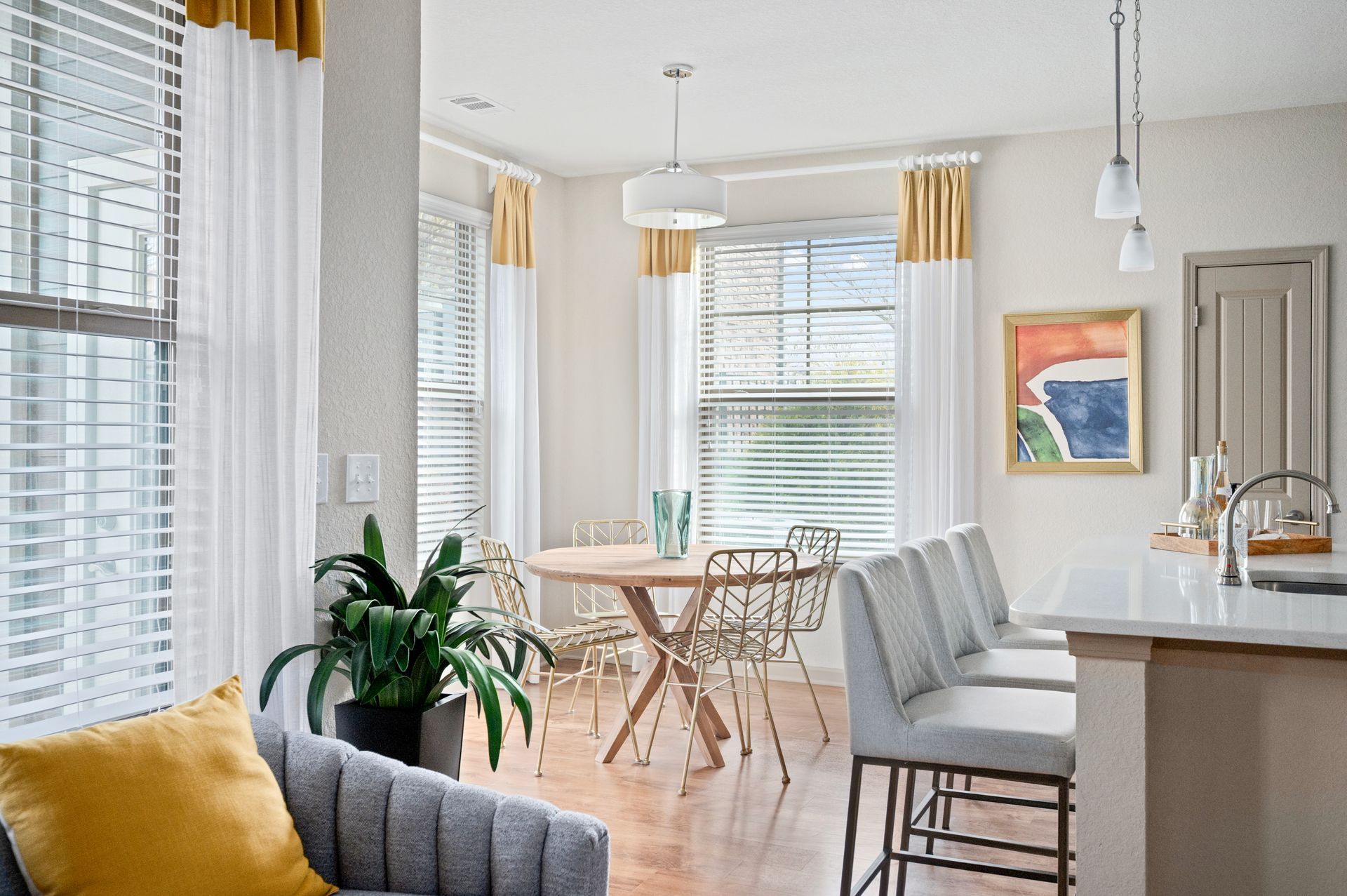 A dining area with a round wood table, metal chairs, hanging pendant lights, and large windows with white and gold drapes.