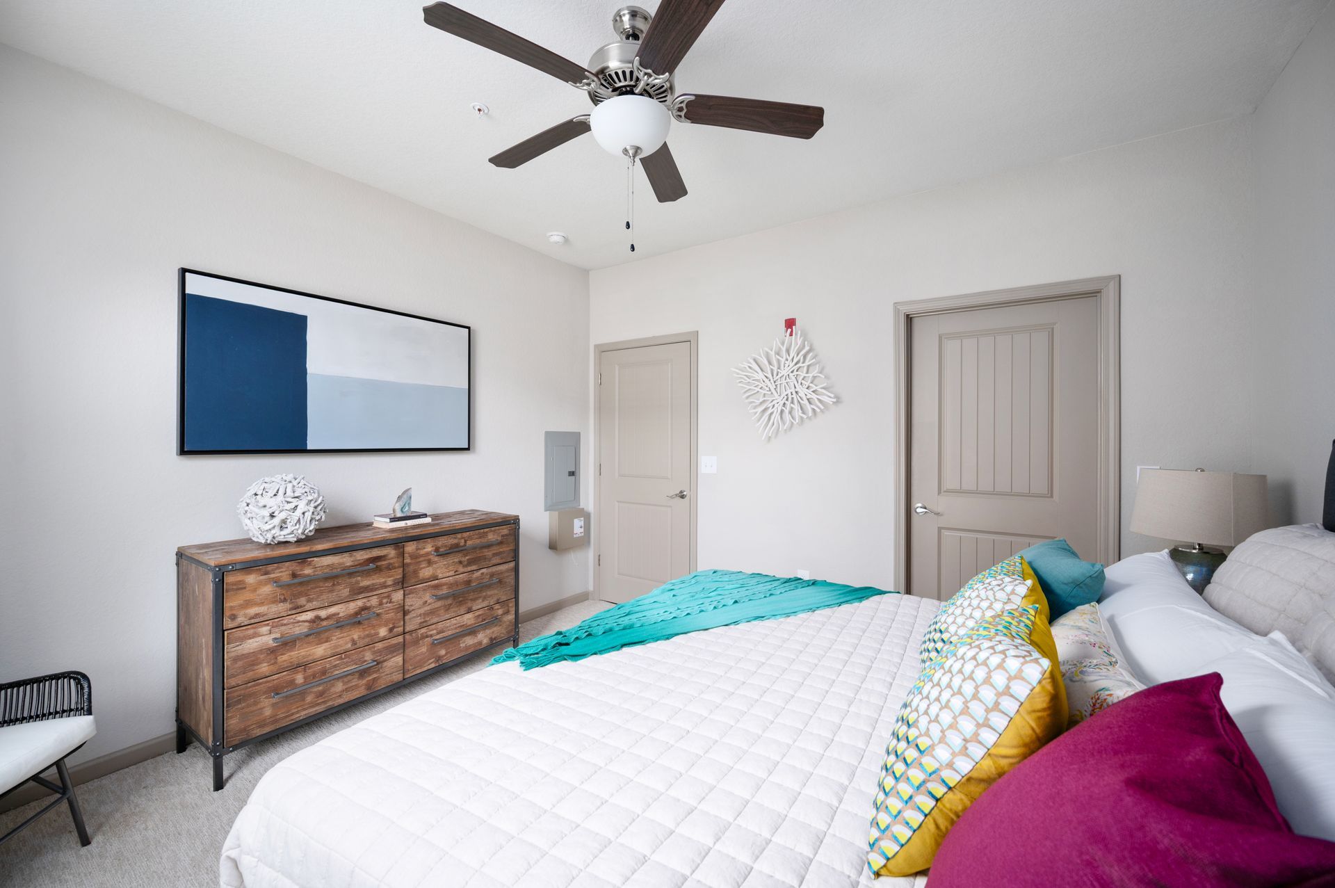 A bright bedroom with a white bed, colorful pillows, a wooden dresser, abstract art, and a ceiling fan at Silver Collection at Cosner's Corner in Fredericksburg, VA.