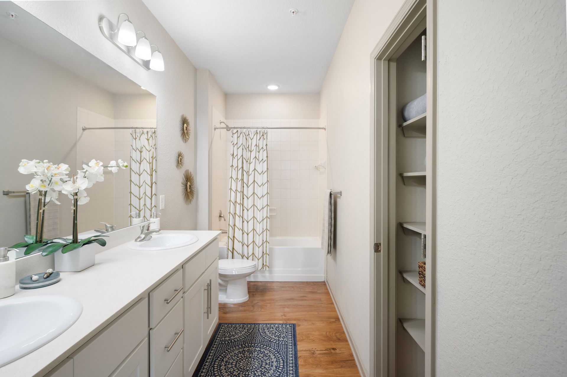 A brightly lit bathroom with a double vanity, a tub-shower combo with a patterned curtain, and open shelving at Silver Collection at Cosner's Corner in Fredericksburg, VA.