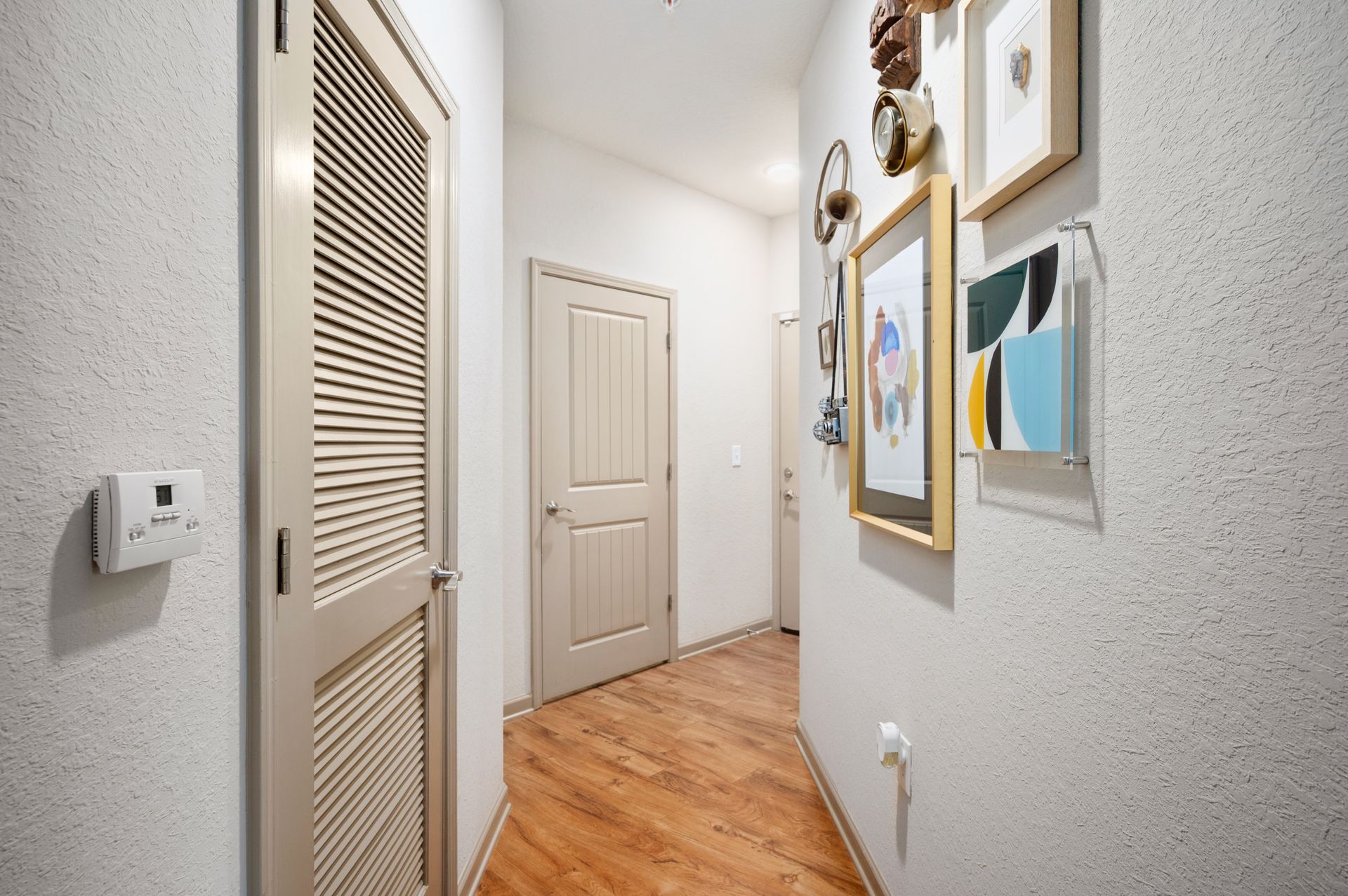A narrow hallway with light wood flooring, beige walls, a louvered door, a standard door, and framed art on the wall at Silver Collection at Cosner's Corner in Fredericksburg, VA.