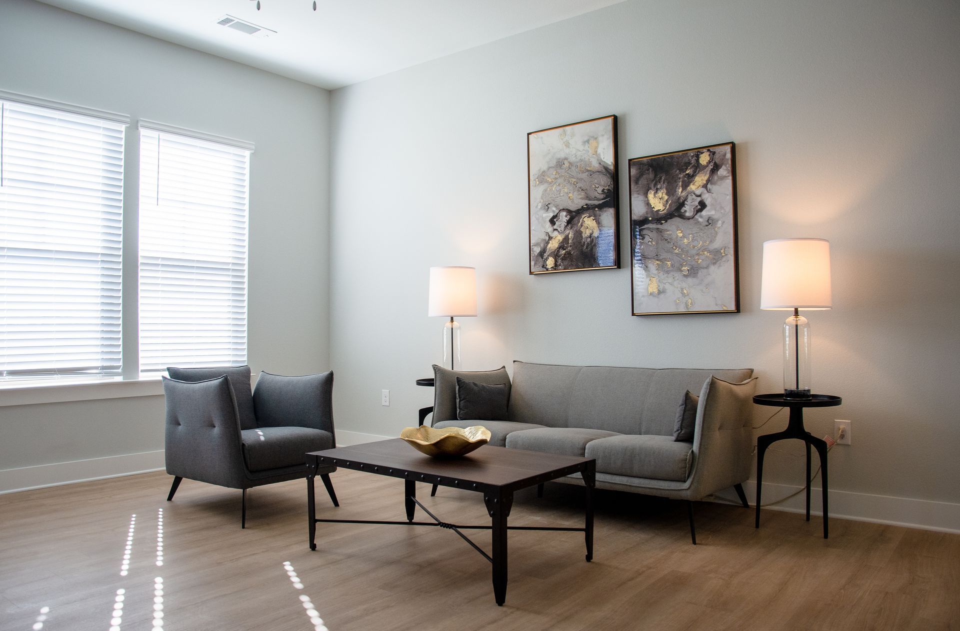 A modern living room with gray furniture, artwork, and lamps. A coffee table sits in front of the couch and chair near a window.