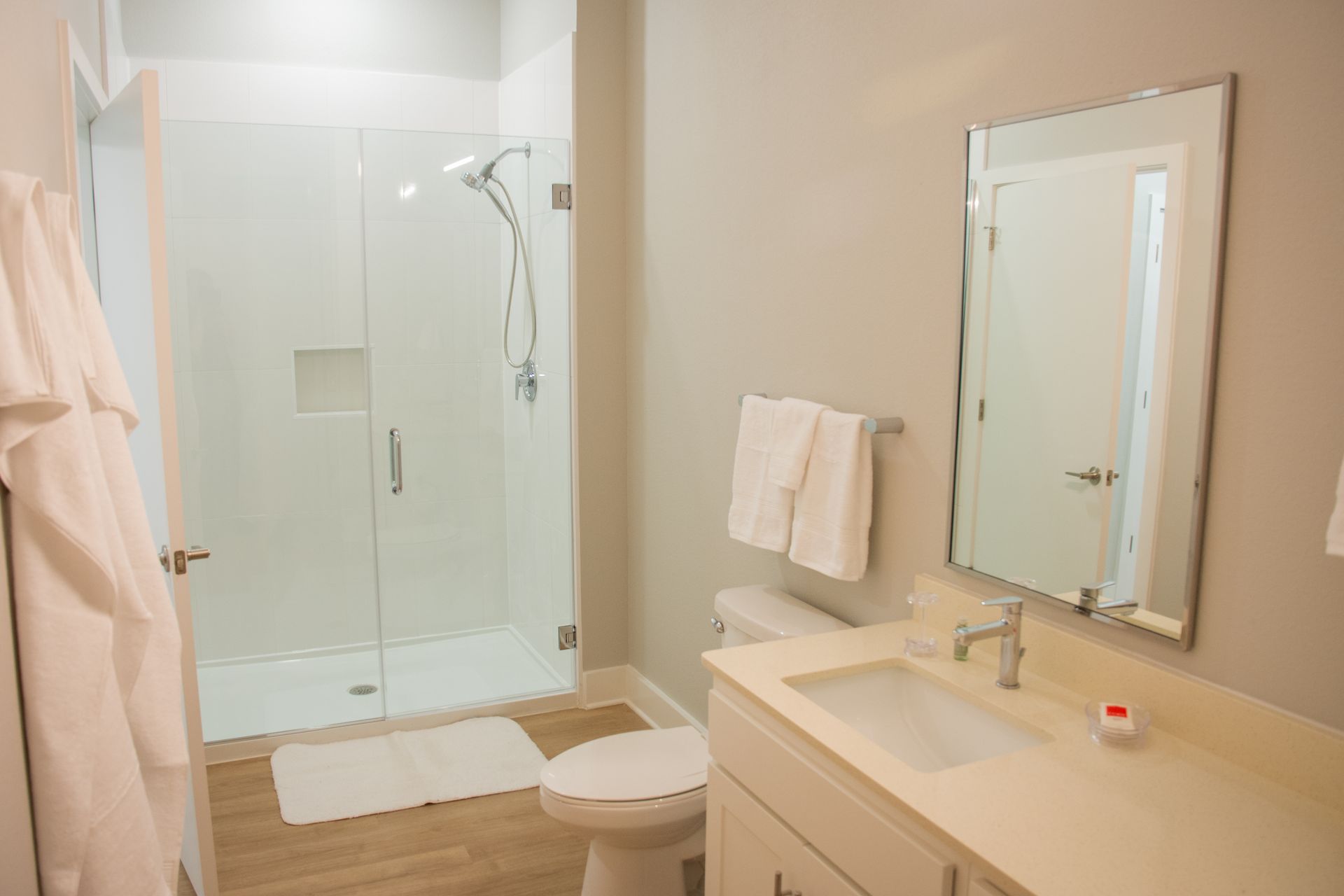 A bright, modern bathroom with a glass shower, white fixtures, and light gray walls. Includes a sink, toilet, mirror, and robe hanging on the door.