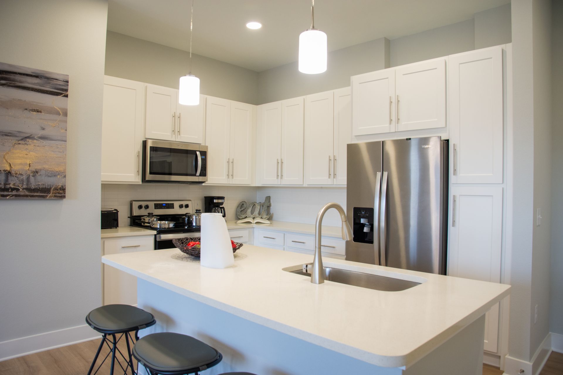 Modern white kitchen with a stainless steel refrigerator, island with sink, and pendant lights.