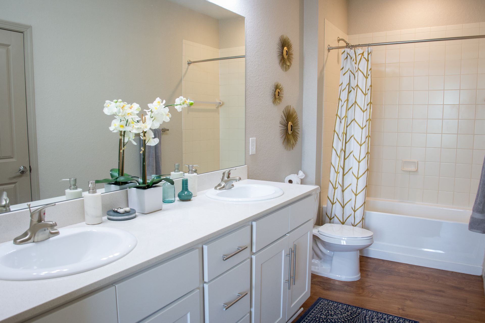 A modern bathroom with white cabinets, double sinks, and a bathtub/shower combo. Decor includes flowers and gold accents.