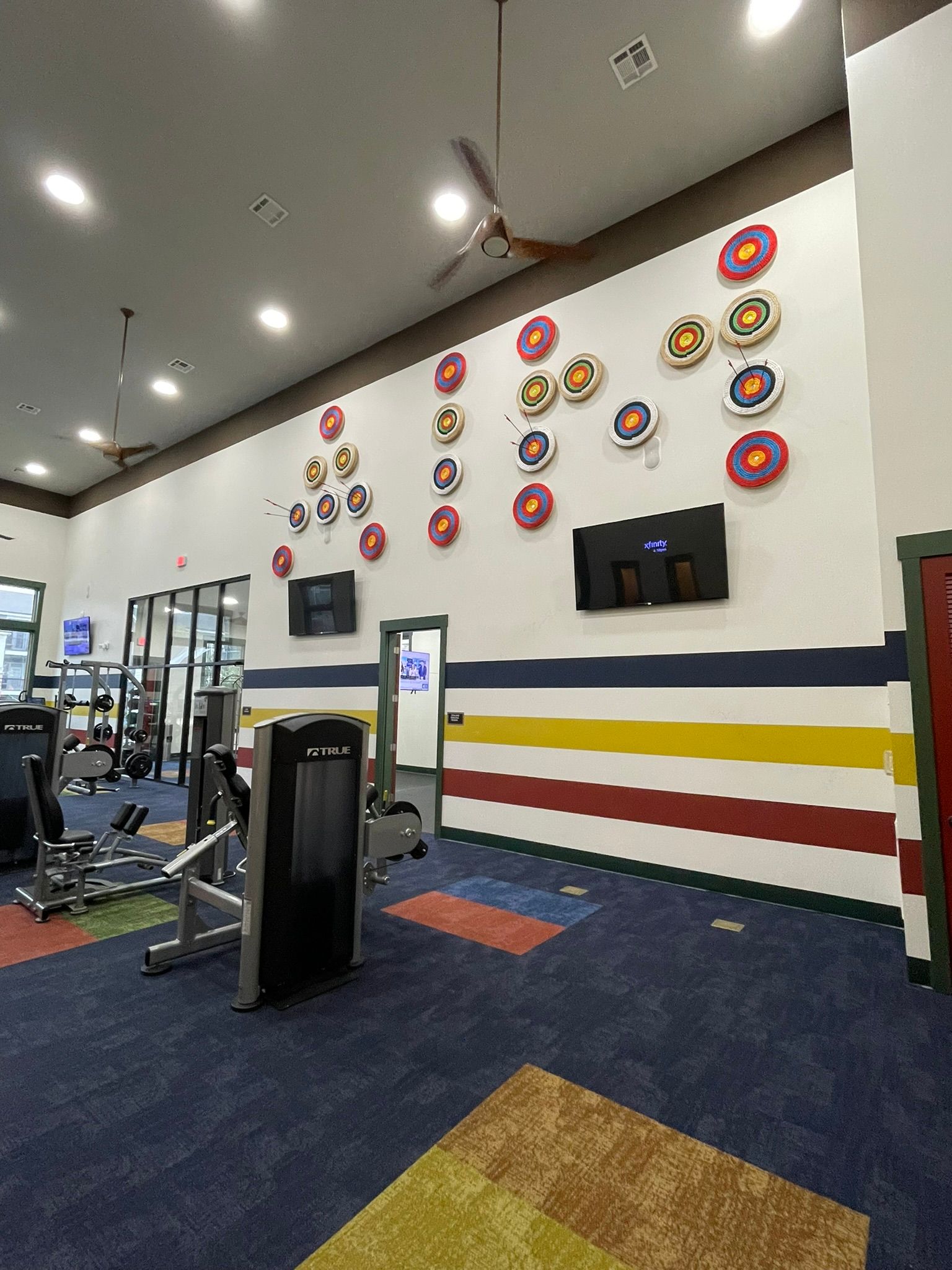 A gym interior featuring a white wall decorated with multiple circular archery targets, exercise equipment, and rug tiles.