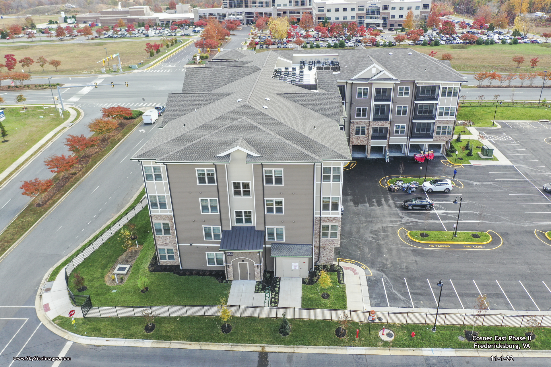 Aerial view of a multi-story apartment building with a gray roof, surrounded by parking lots and roads in a suburban setting.