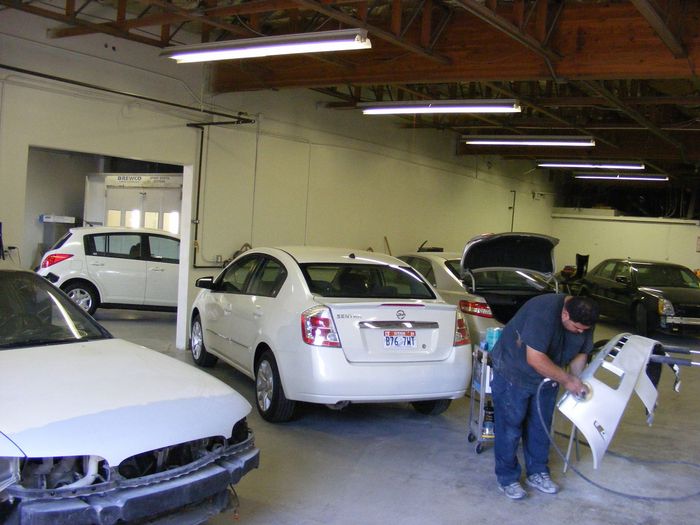Auto body shop with several cars and a worker repairing a white bumper indoors