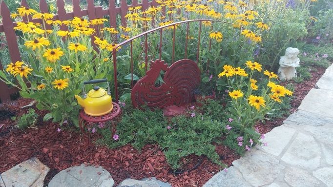 A garden with yellow flowers , a tea kettle , and a statue.