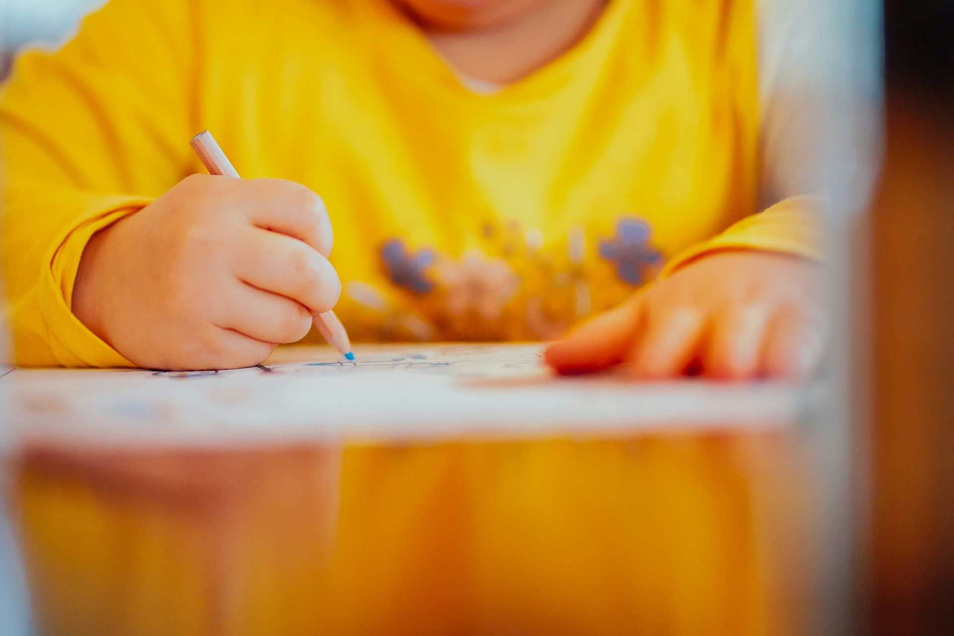 Child in a yellow shirt writing with a pencil on paper at a table
