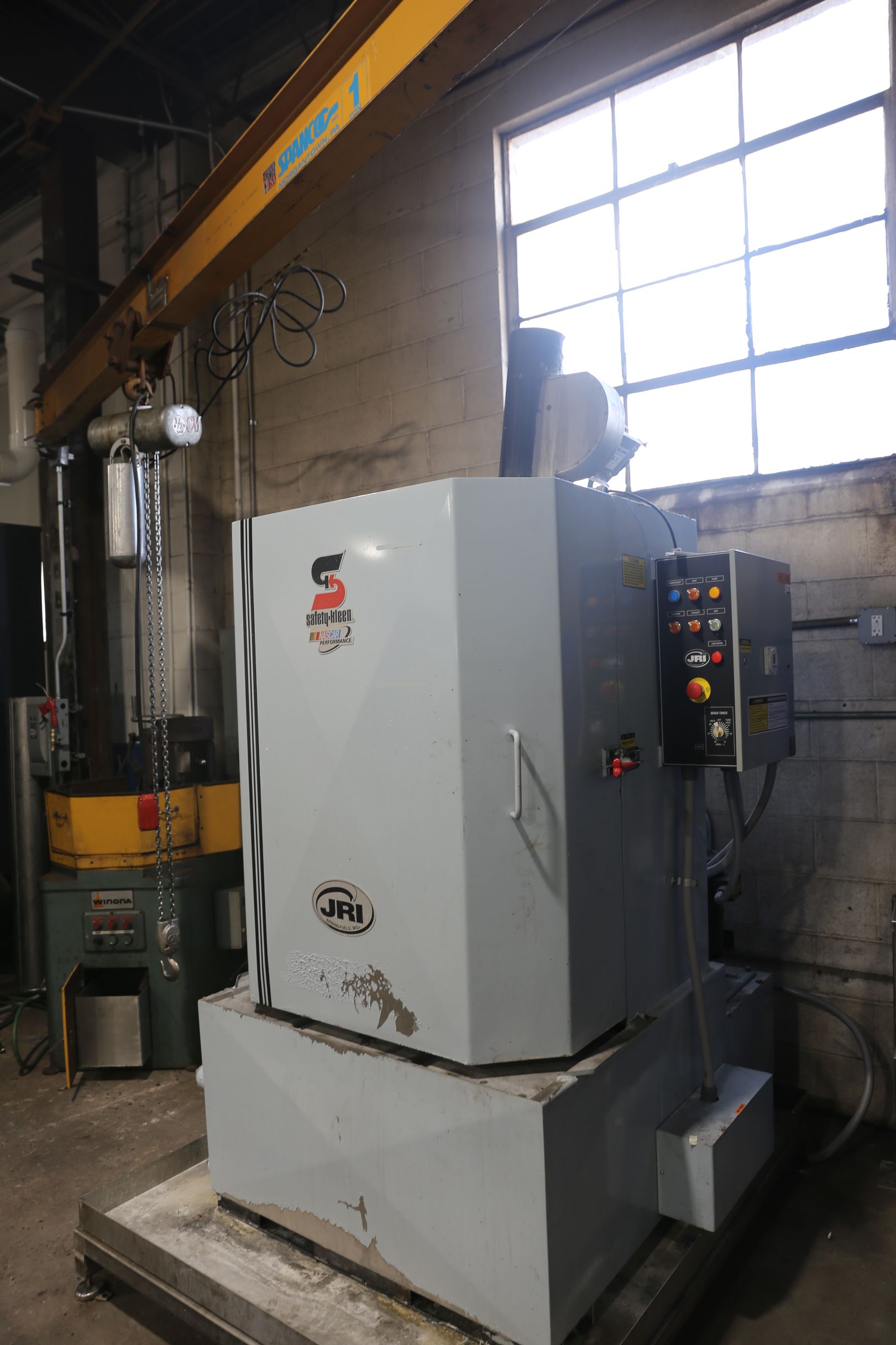 Gray industrial machine with control panel, under a yellow crane, next to a window.