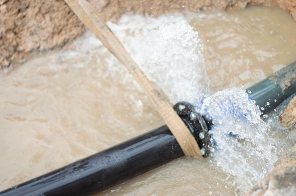Black pipe bursts with water spraying into a muddy trench