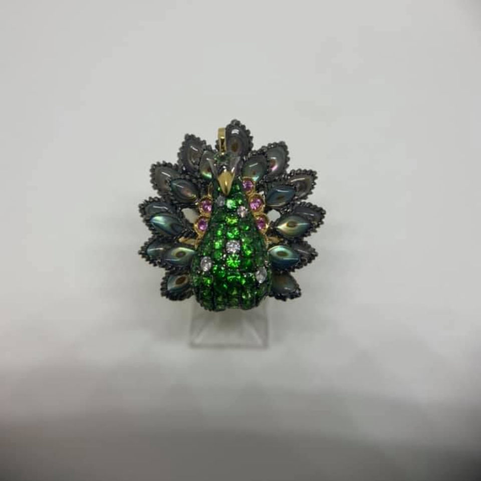 Jeweled peacock brooch with green and multicolored stones on a clear stand against a gray background