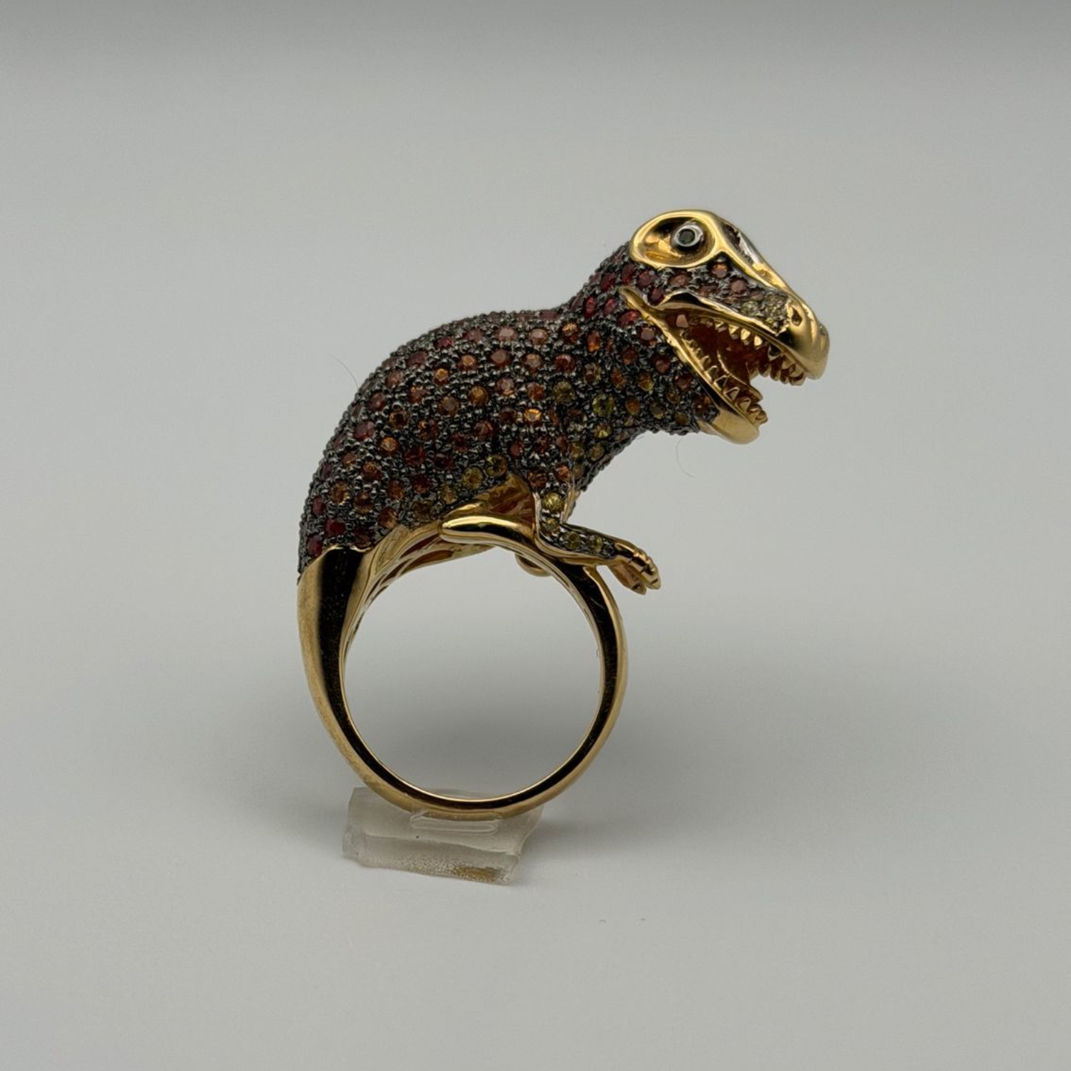 Gold-and-black dinosaur-shaped ring on a clear stand against a gray background