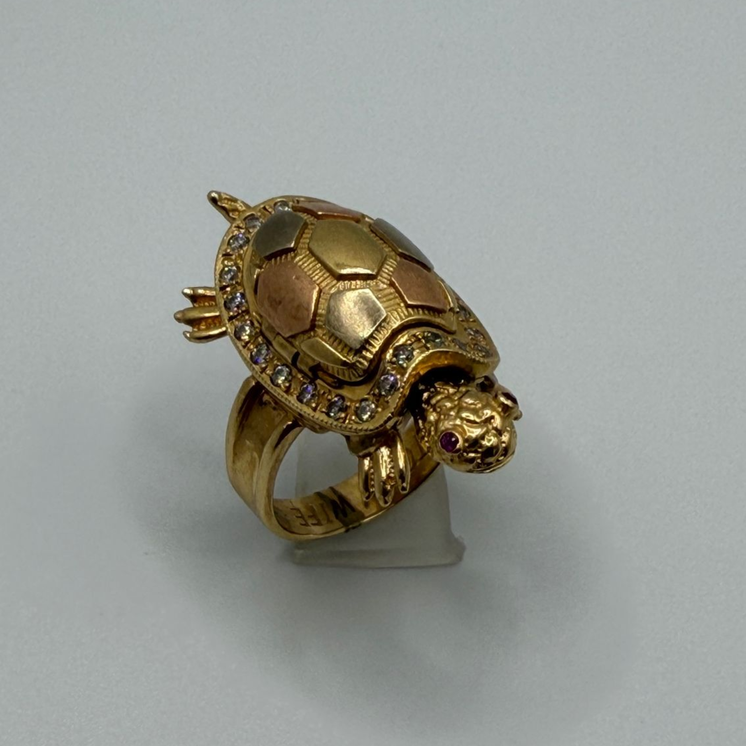 Gold turtle-shaped ring on a stand against a plain gray background
