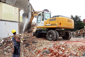 Una excavadora demuele un edificio de ladrillos mientras un trabajador rocía agua