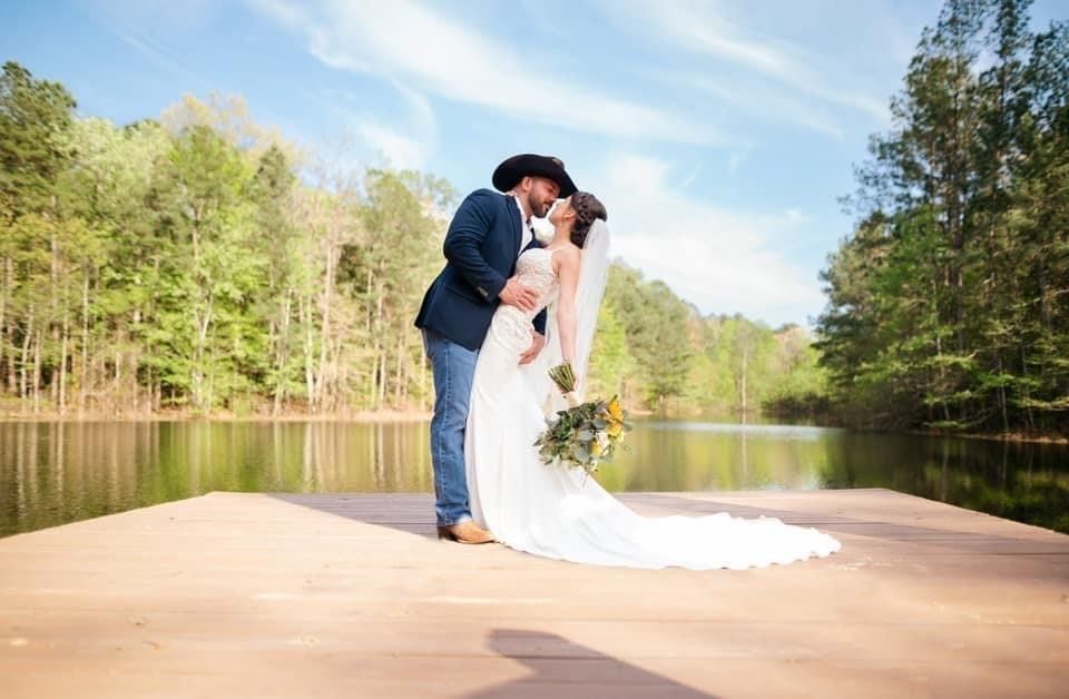 Bride and groom kiss on a wooden dock overlooking a lake. The man wears a cowboy hat and the woman holds a bouquet.
