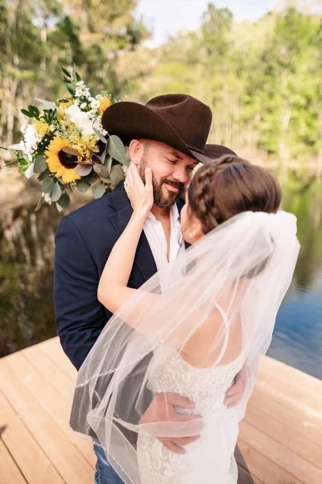 Bride and groom embrace on a wooden dock; groom in cowboy hat, bride in veil, holding bouquet.