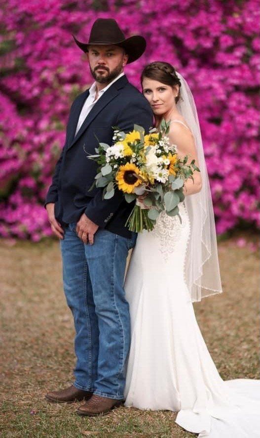 Wedding couple posing in front of pink flowers; man in cowboy hat and suit jacket, woman in white dress holding a bouquet.
